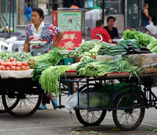 中国の市場で女性がリヤカーに乗って野菜を売っている。