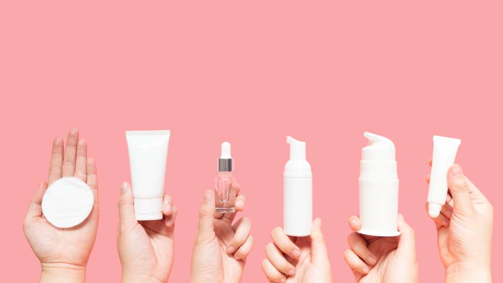 ピンクの背景にさまざまな化粧品を持つ女性の手。
