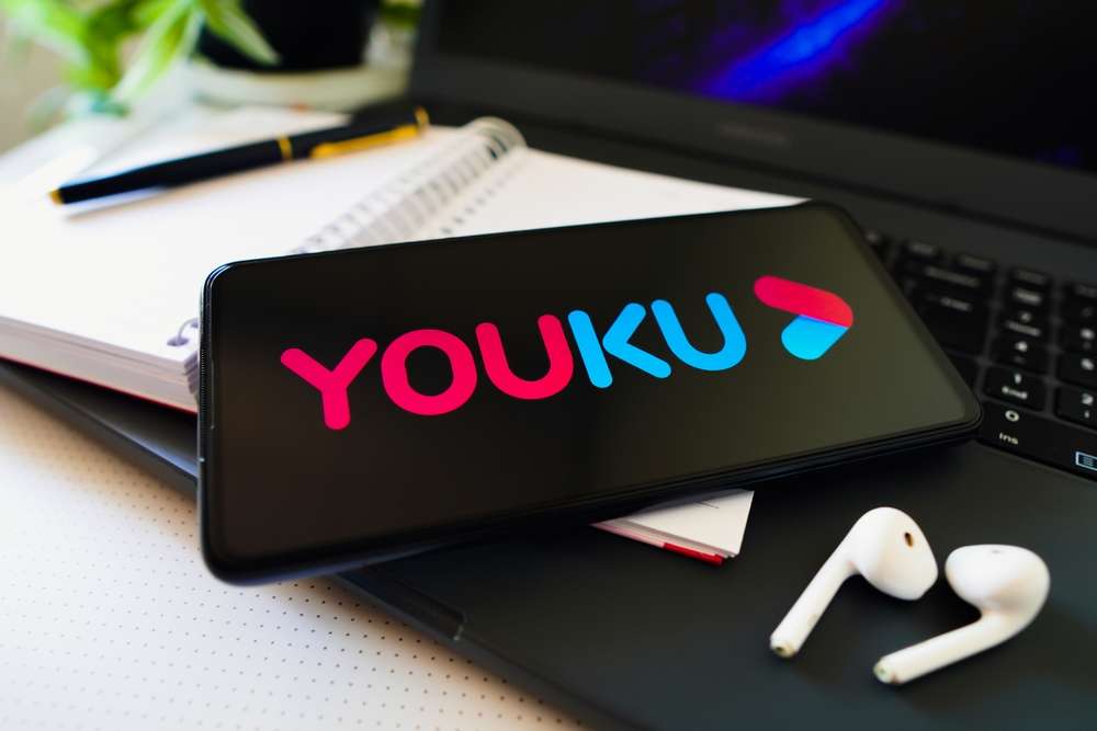 Youku のロゴが表示されたスマートフォンが、白いイヤホン、ノート、ペンの横にあるノートパソコンのキーボードの上に置かれています。これは、中国のトップ ストリーミング コンテンツを楽しむための理想的なセットアップです。