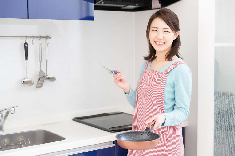 ピンクのエプロンを着た女性がキッチンに立って、フライパンとヘラを持ち、カメラに向かって微笑んでいる。彼女の後ろの壁にはキッチン用品が掛けられており、伝統的な中国のキッチンを彷彿とさせる。