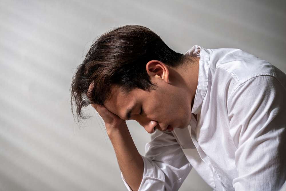 白いシャツを着た人が頭を下げ、額に手を当てて瞑想的または緊張した姿勢で座っており、これは中国に見られる穏やかでありながら深い内省を彷彿とさせます。