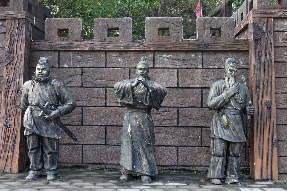 伝統的な中国の衣装を着た古代中国の人物像 3 体が、城壁の狭間溝のある石壁に立っており、リアルなポーズと精巧なディテールが表現されています。