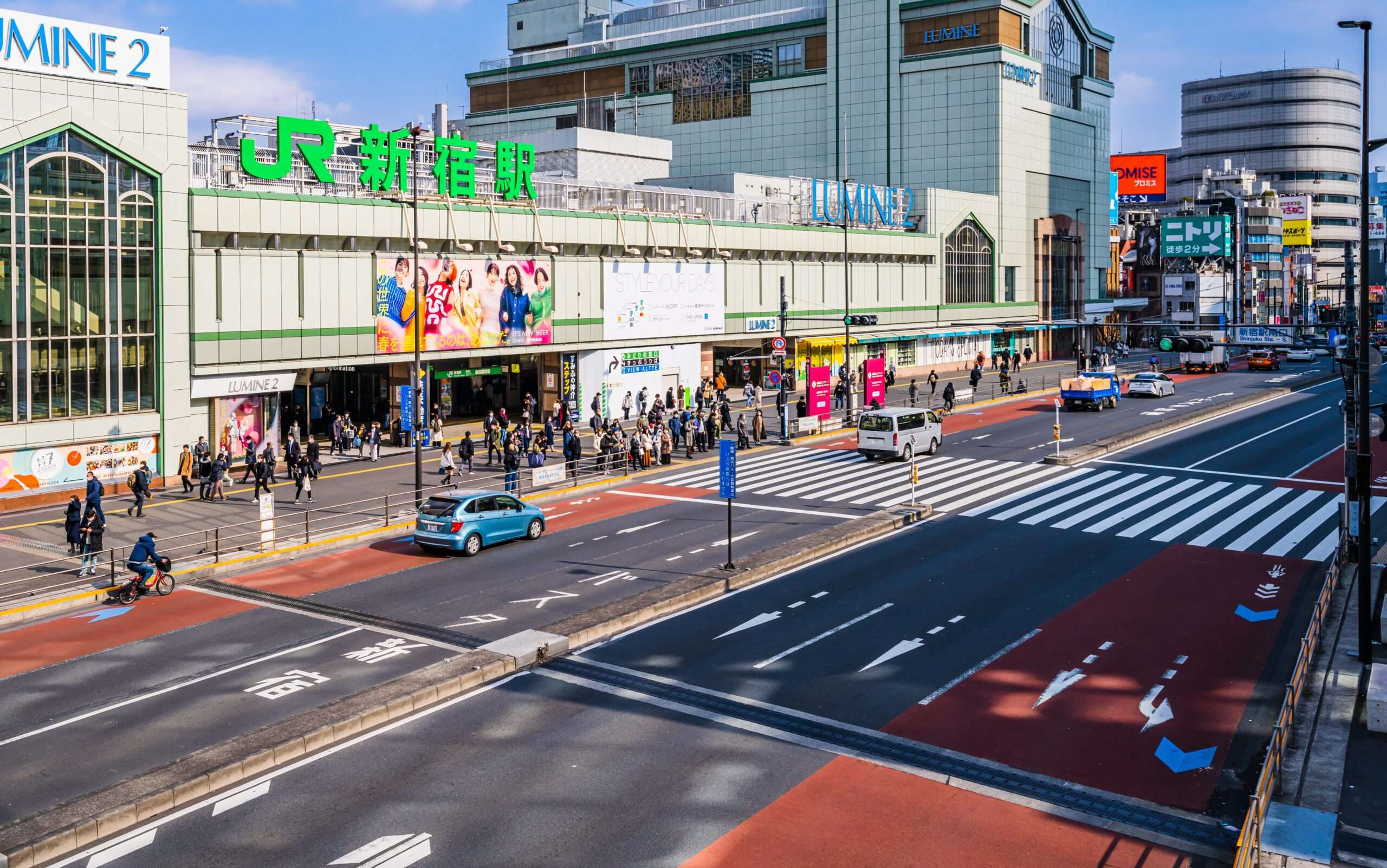 東京のJR新橋駅前の賑やかな通りには、歩行者や車、そして鮮やかな看板が並んでいます。活気ある風景の中に、中国語教室を指し示す看板があり、この地域の文化的な雰囲気を一層引き立てています。
