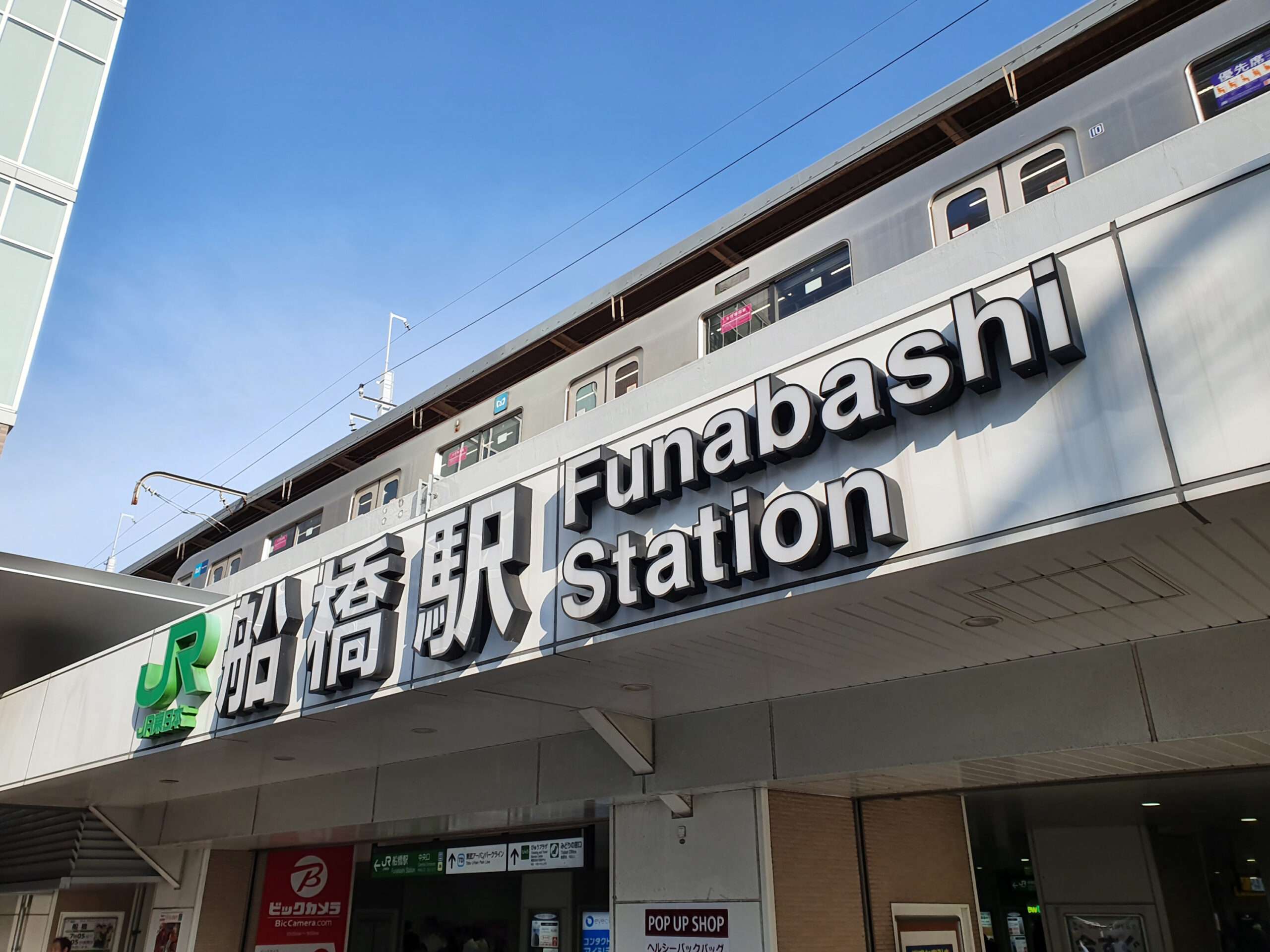 日本の船橋駅の外観。日本語と英語の両方で目立つ標識があり、上の高架線には電車が見えます。船橋周辺の賑やかなエリアには、熱心な学習者のための中国語教室などの語学機関もあります。