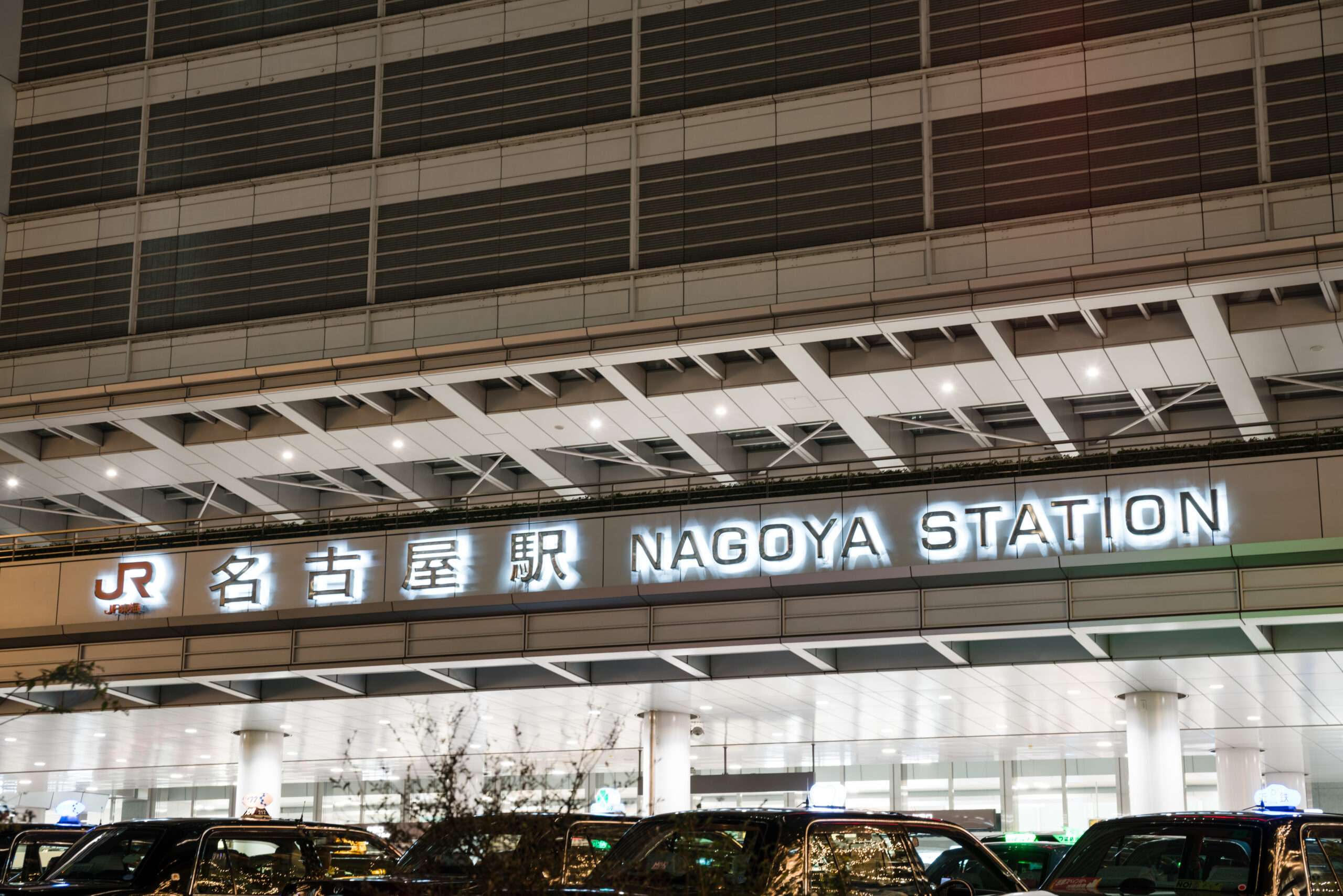 夜にライトアップされた名古屋駅の駅名標の正面図。太字の英語と日本語の文字が目立ち、その下には車が駐車している。