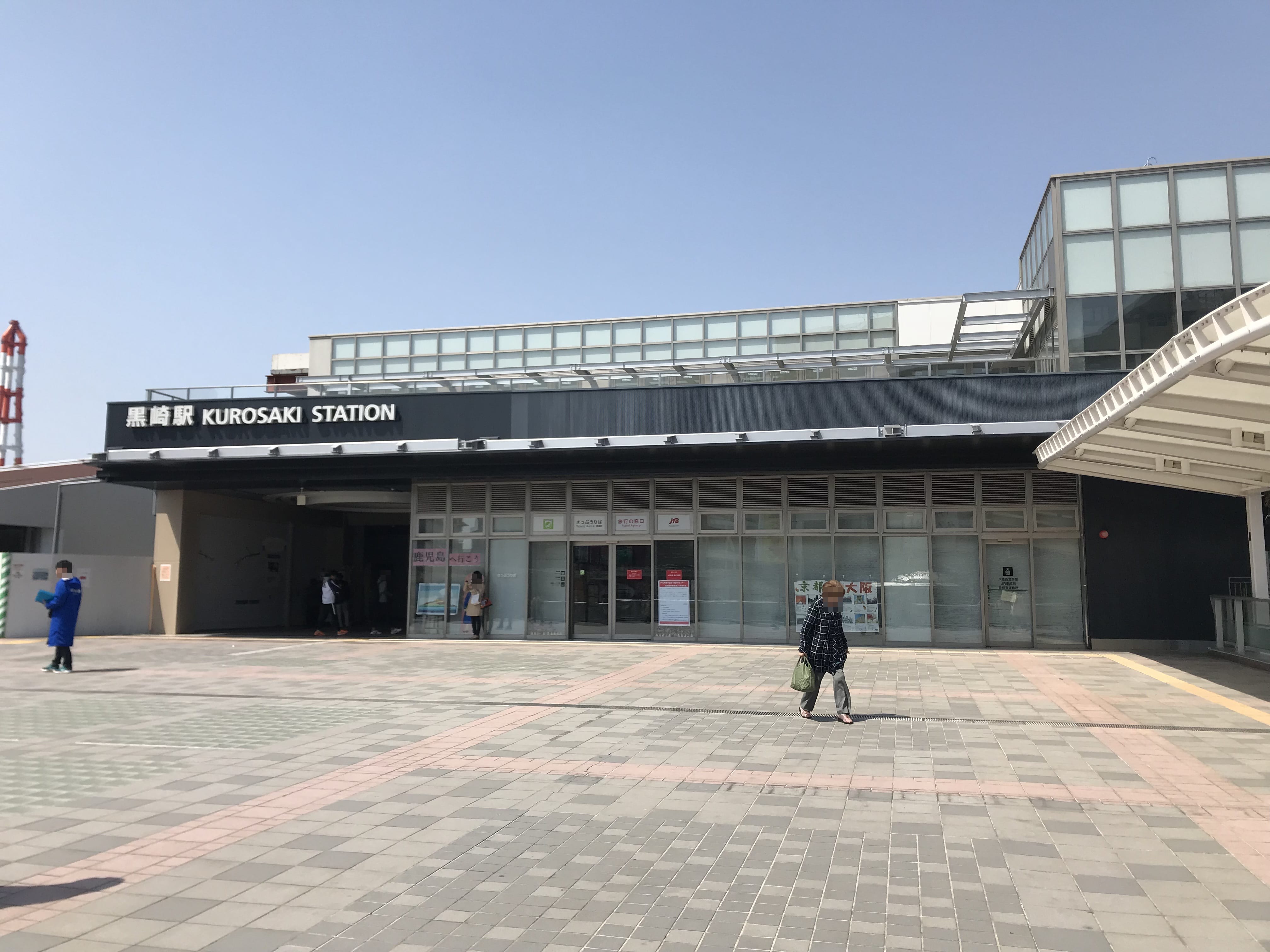 晴れた日の黒崎駅外観。ガラス扉、日本語と英語の看板、広場を歩く数人の人々。.