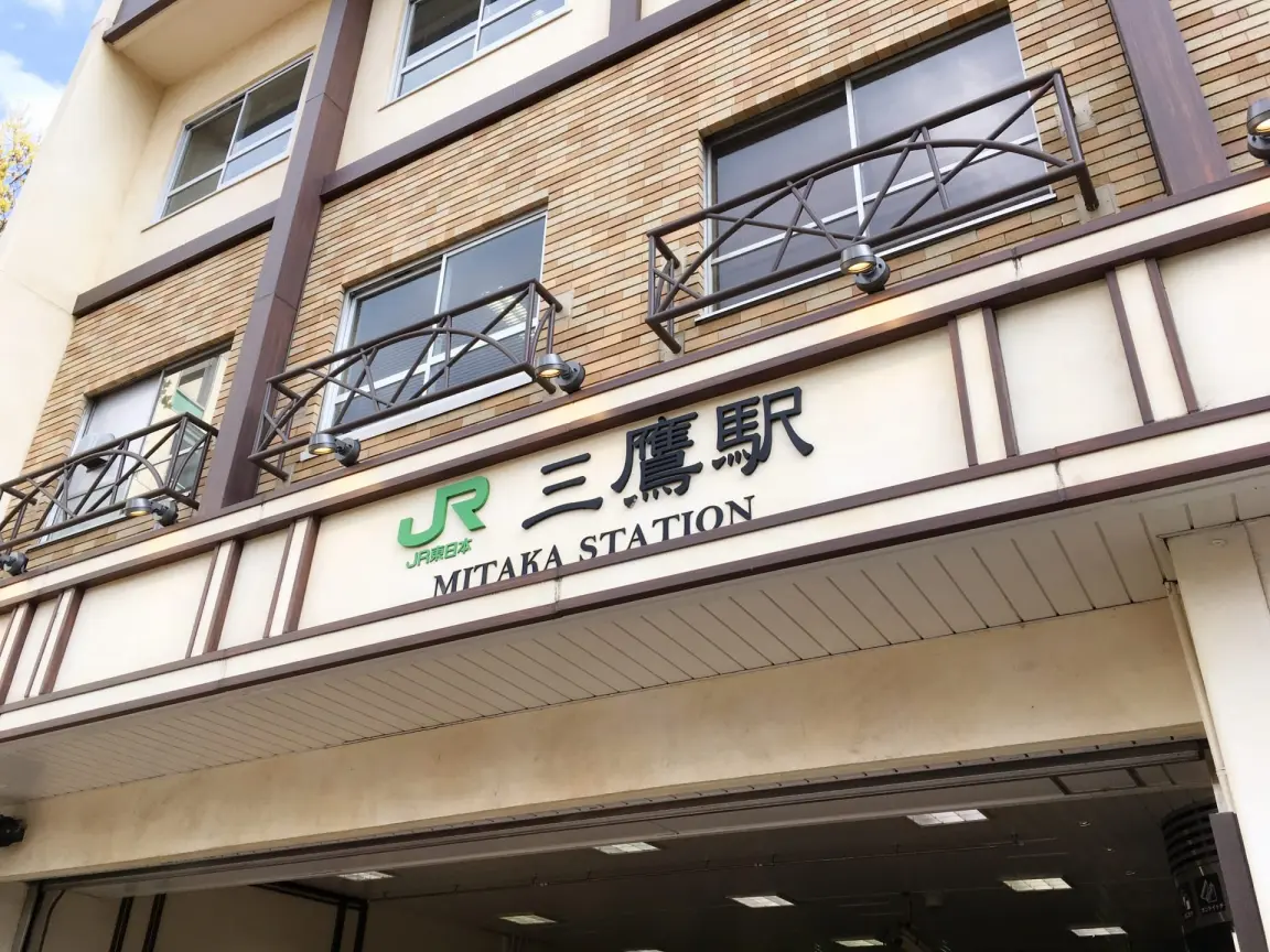日本の三鷹駅のエントランス。JRのロゴと、ビルのファサードにある日本語と英語の看板が見える。.