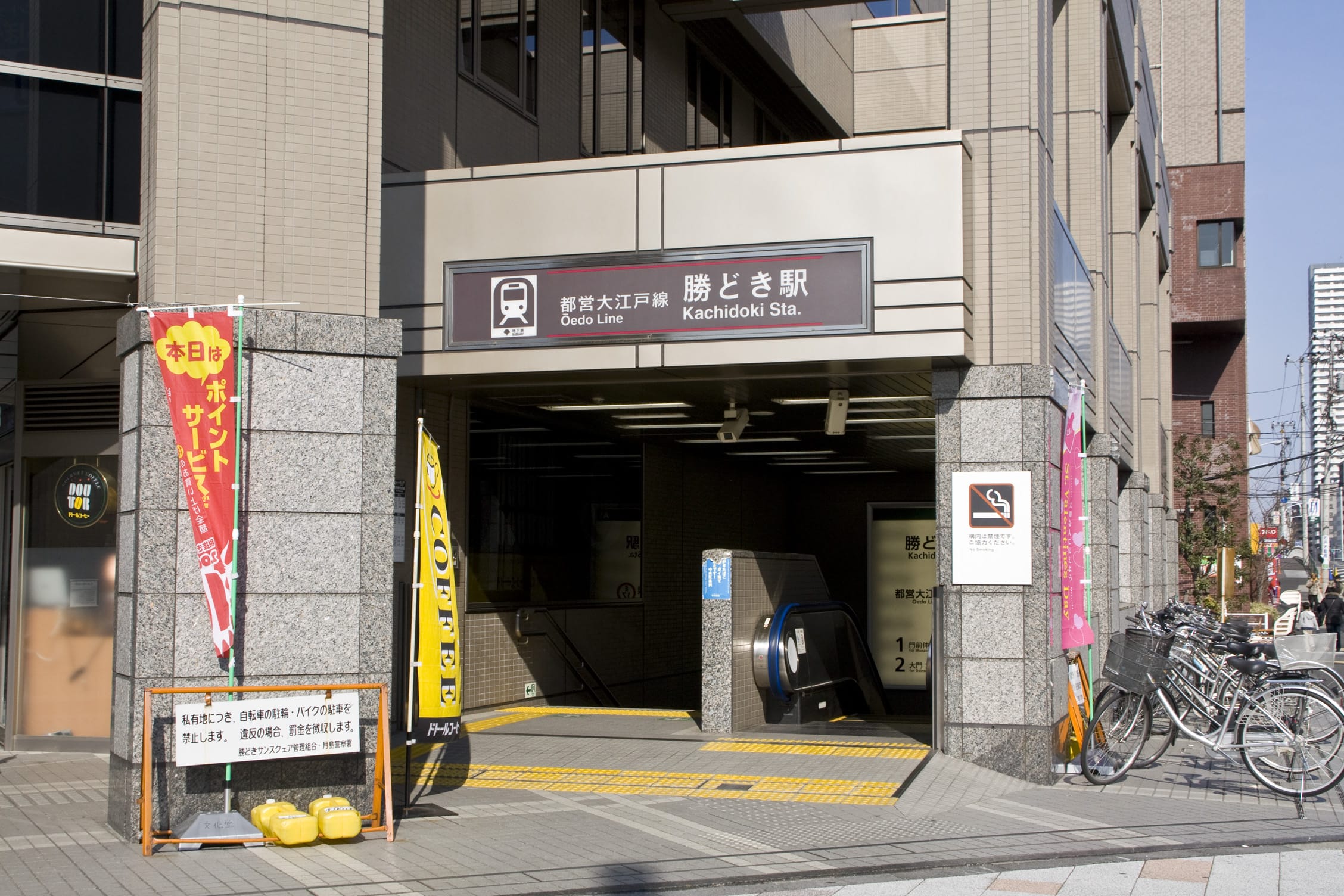 都営大江戸線勝どき駅入り口。エスカレーター、日本語の看板、自転車、カラフルな横断幕が近くにある。.