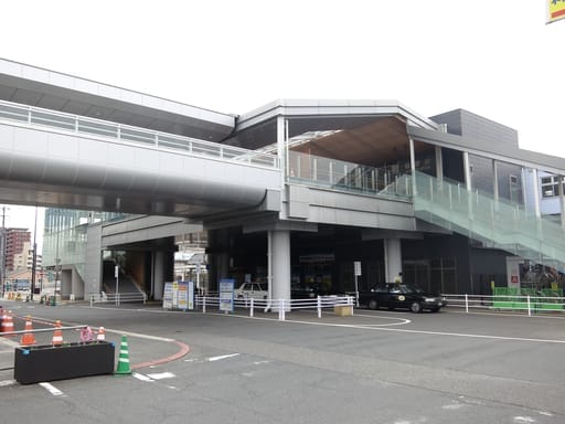 高架の歩行者用通路、ガラスの手すり、入り口に停まるタクシーを備えた近代的な立体駅舎。.