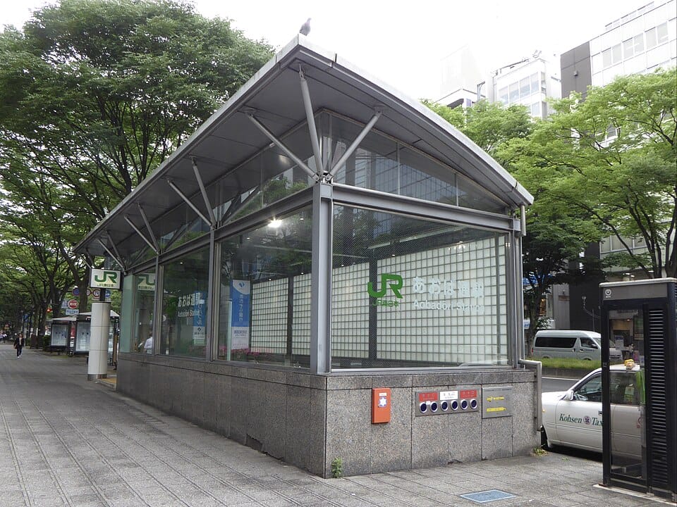 モダンなガラス張りのJR駅の入り口。仙台駅の看板があり、木々や街の建物に囲まれている。.