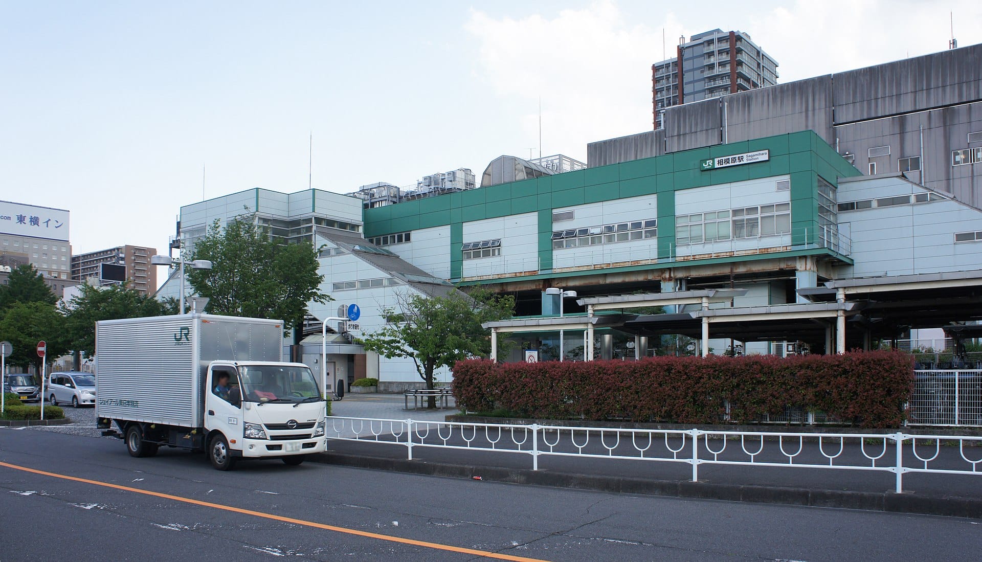 緑と白の大きなビルの前に配送トラックが路上駐車しており、背景には木々と高層ビルがある。.