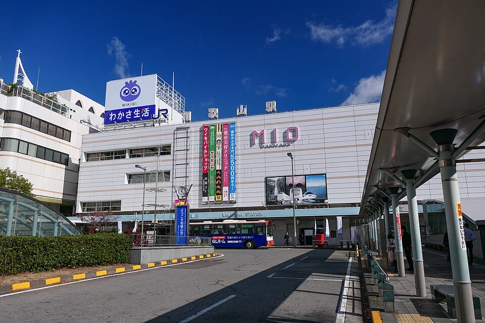 快晴の青空の下、バス停エリア、屋根付き通路、日本語と英語の標識があるJR和歌山駅の外観。.