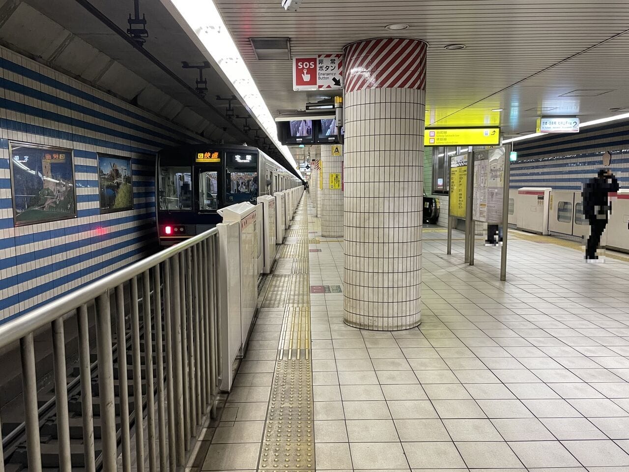 壁と床がタイル張りの地下駅に停車した地下鉄。ホームを歩く人が1人、頭上には標識が見える。.