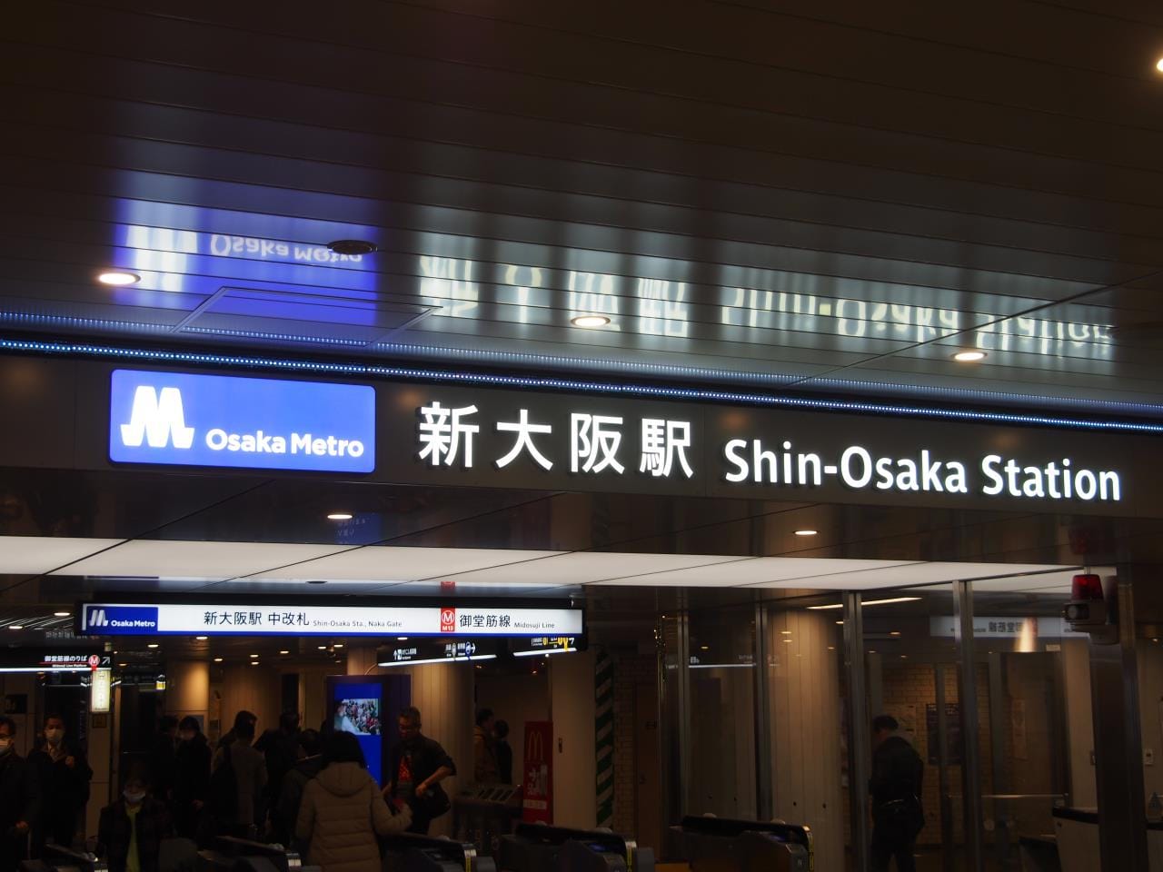 大阪メトロのロゴ、日本語と英語の電飾看板、改札口付近の人影がある新大阪駅入口。.