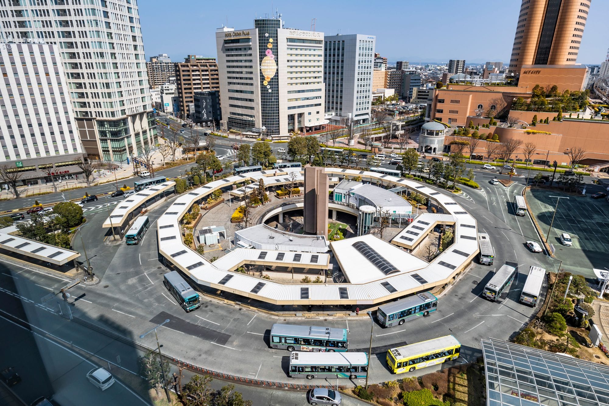 市街地の建物に囲まれた円形のバスターミナルの上空からの眺め。晴れた日には駅周辺に数台のバスが停車し、走っている。.