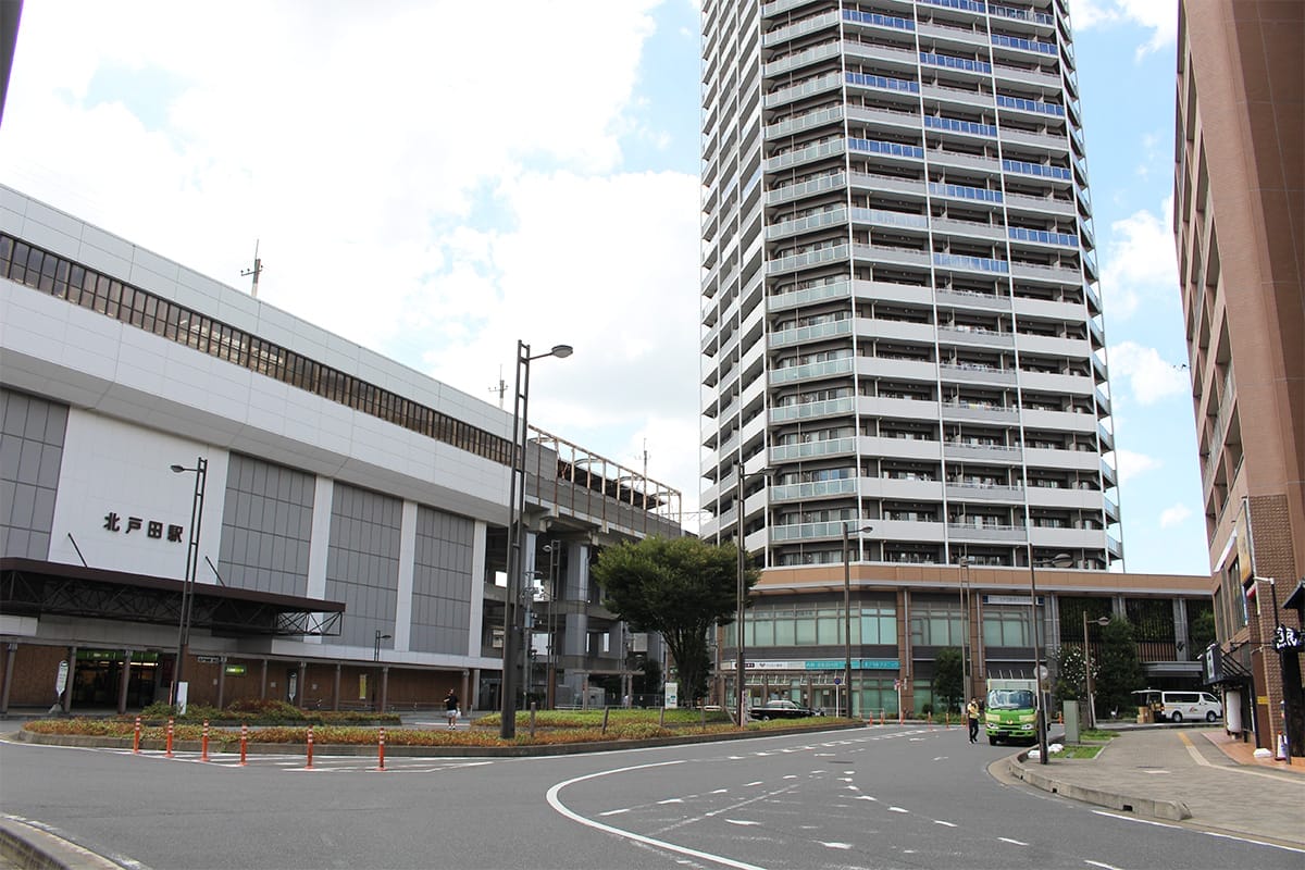 一部曇り空の下、背の高い近代的なマンション、「北戸田駅」と書かれた多層階の駅ビル、そして数台の車が近くに駐車している街並み。.