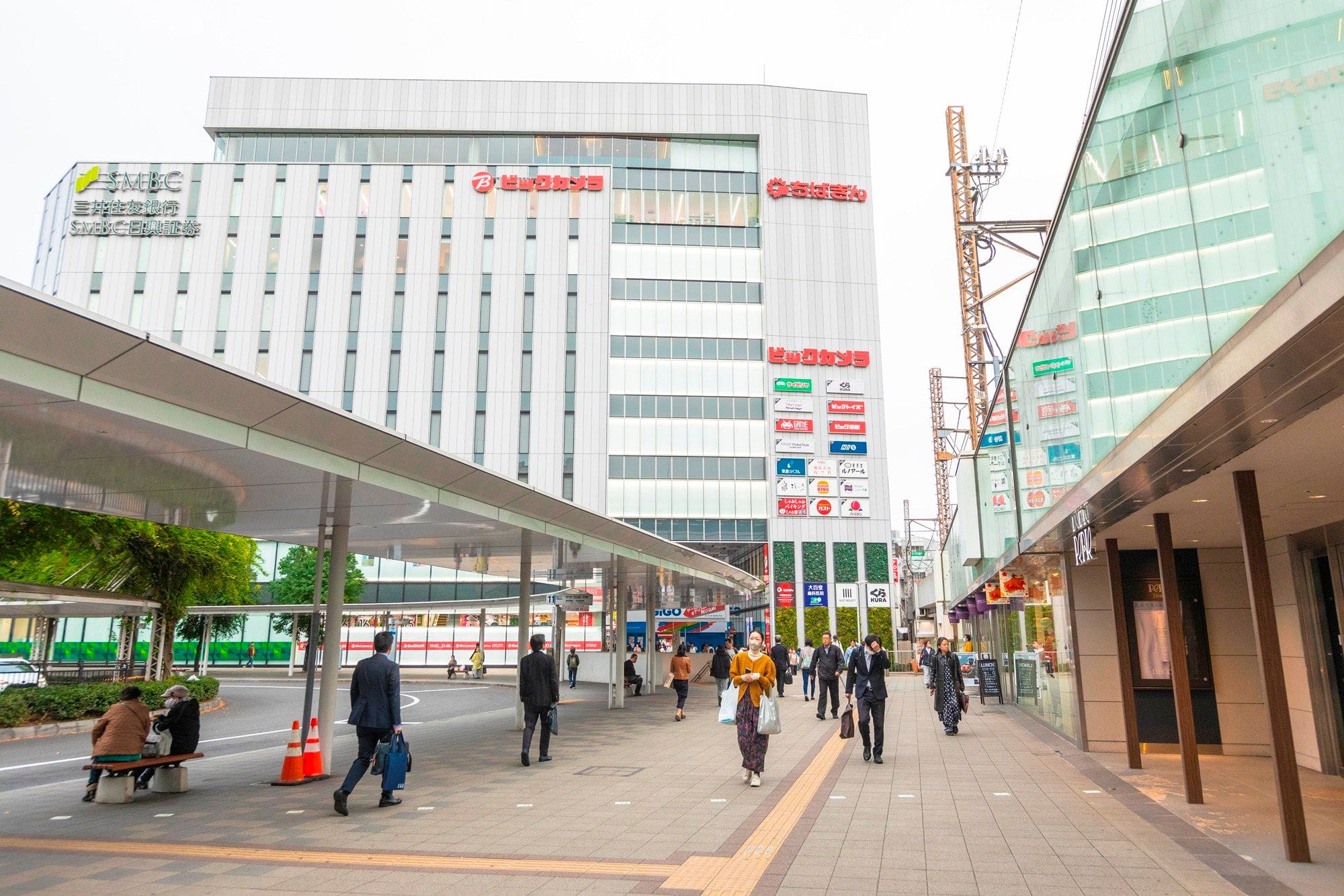 日本の看板が掲げられた近代的なビルに挟まれた広場で、人々は屋根のある歩道や交通量の少ない道路の近くを歩いている。.