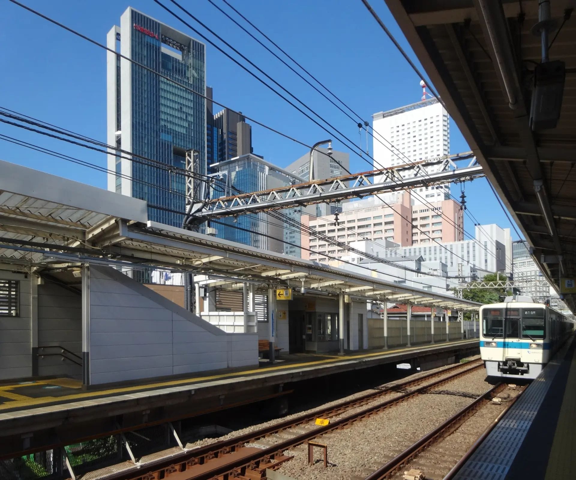 澄み切った青空の下、近代的な高層ビルに囲まれ、線路と列車が近づいてくる都会の駅のホーム。.