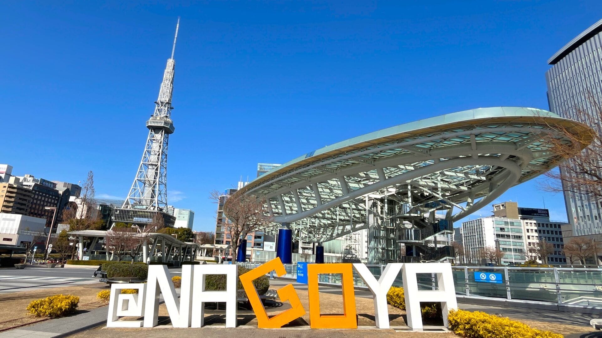 晴れ渡った青空の下、大きな「NAGOYA」の看板、近代的なオアシス21の建造物、名古屋テレビ塔が特徴的な名古屋の屋外広場。.