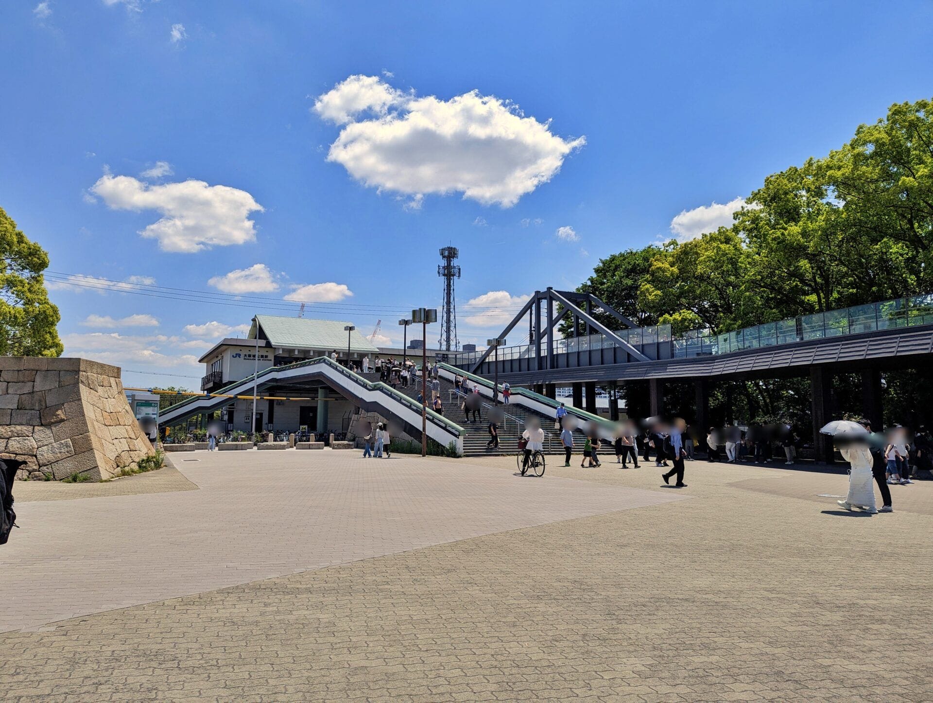 舗装されたオープンエリアで、階段や歩道橋の近くを人々が歩いている。.