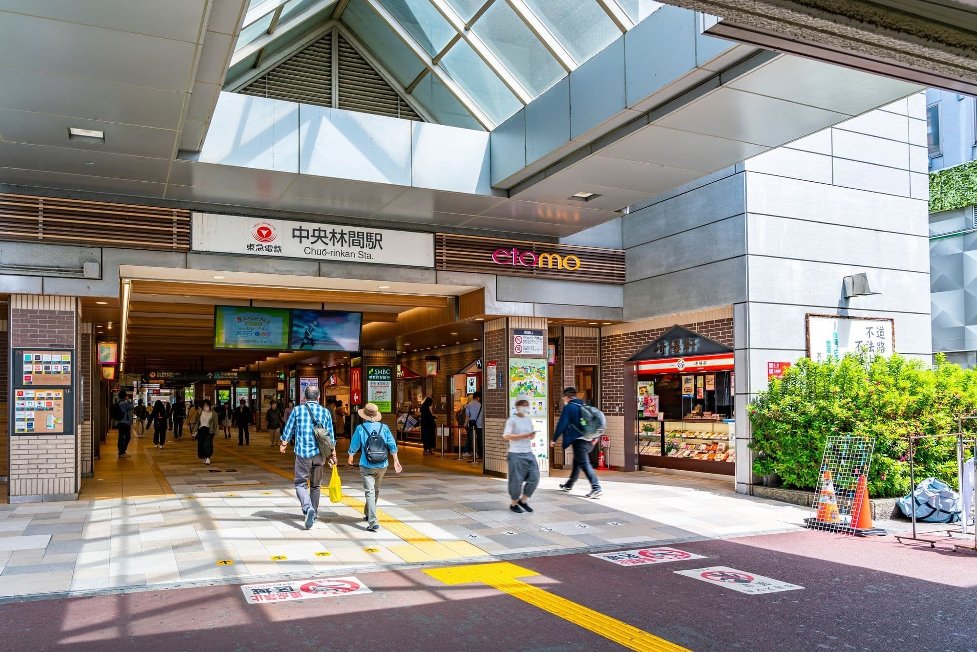 日本の近代的な駅である中央林間駅に出入りする歩行者。入り口付近には店舗や看板が見える。.