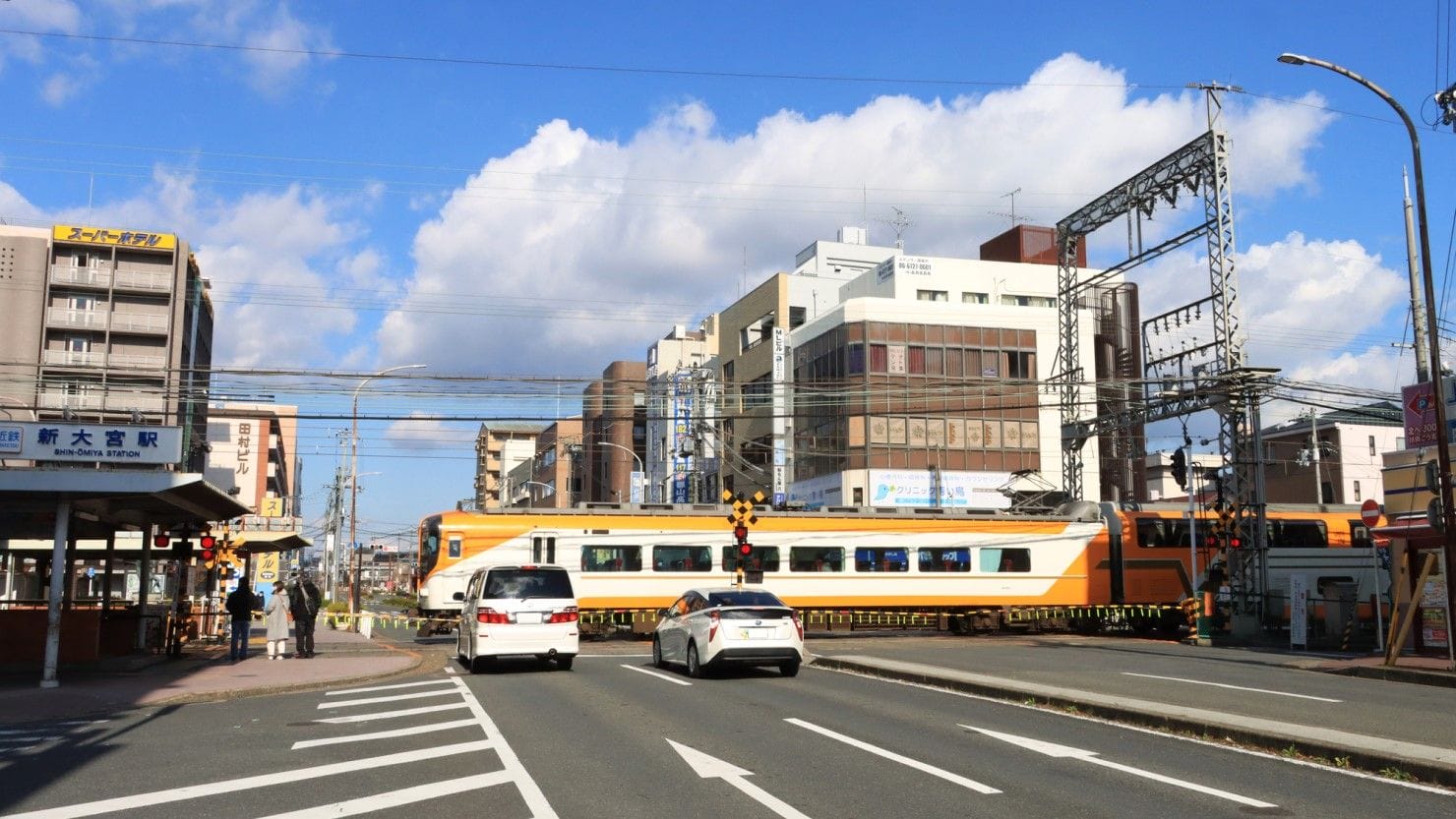 一部曇り空の下、オレンジと白の列車が、待機する車と建物を背景に、市街地の踏切を通過する。.