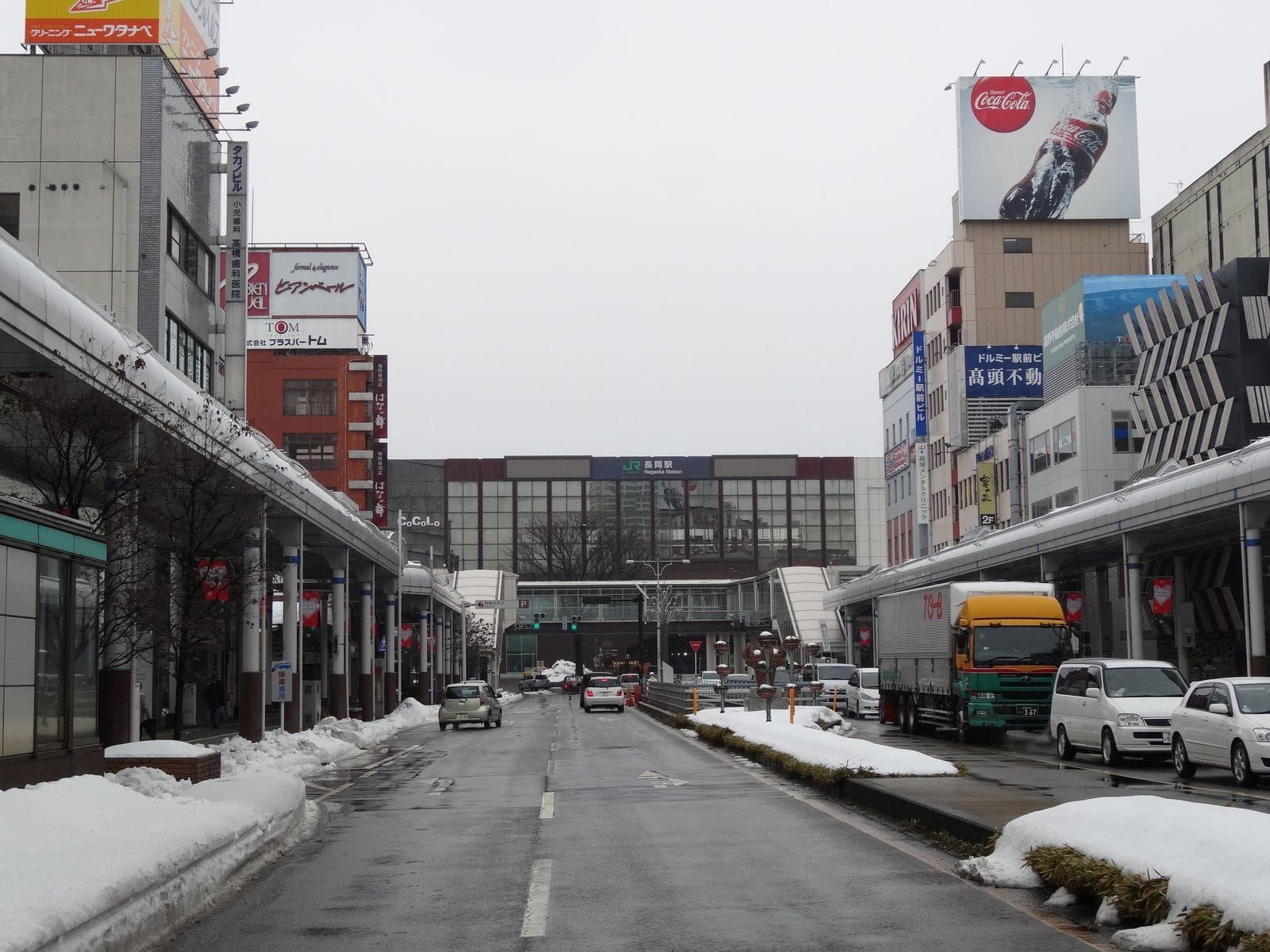 曇り空の下、雪が積もり、ビルや車、トラックが立ち並ぶ市街地。道の端に駅が見える。.