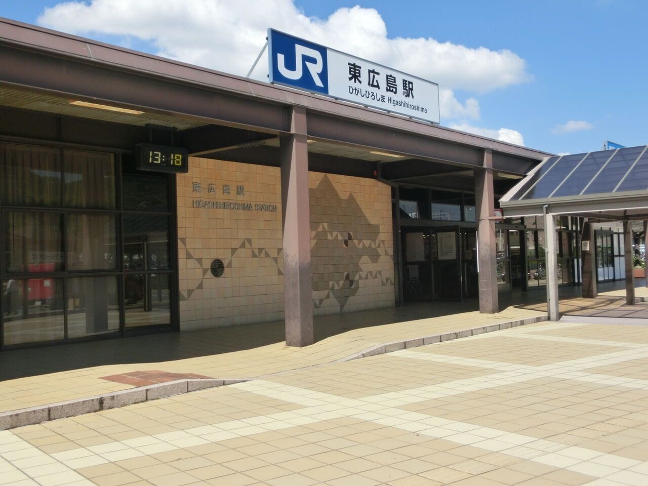 日本の東広島駅の外観。エントランス、タイル張りの通路、13時18分を示すデジタル時計。.