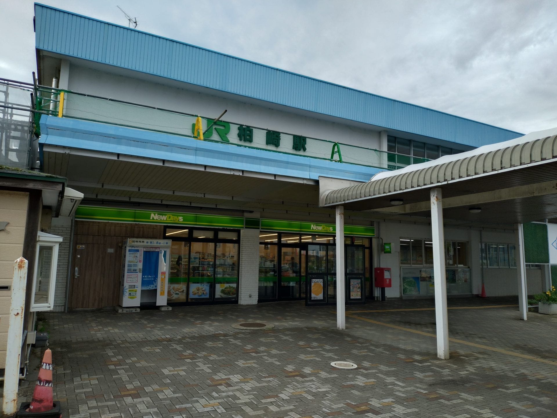 曇り空の下、緑の看板の下にコンビニエンスストア「NewDays」がある駅の入り口。.