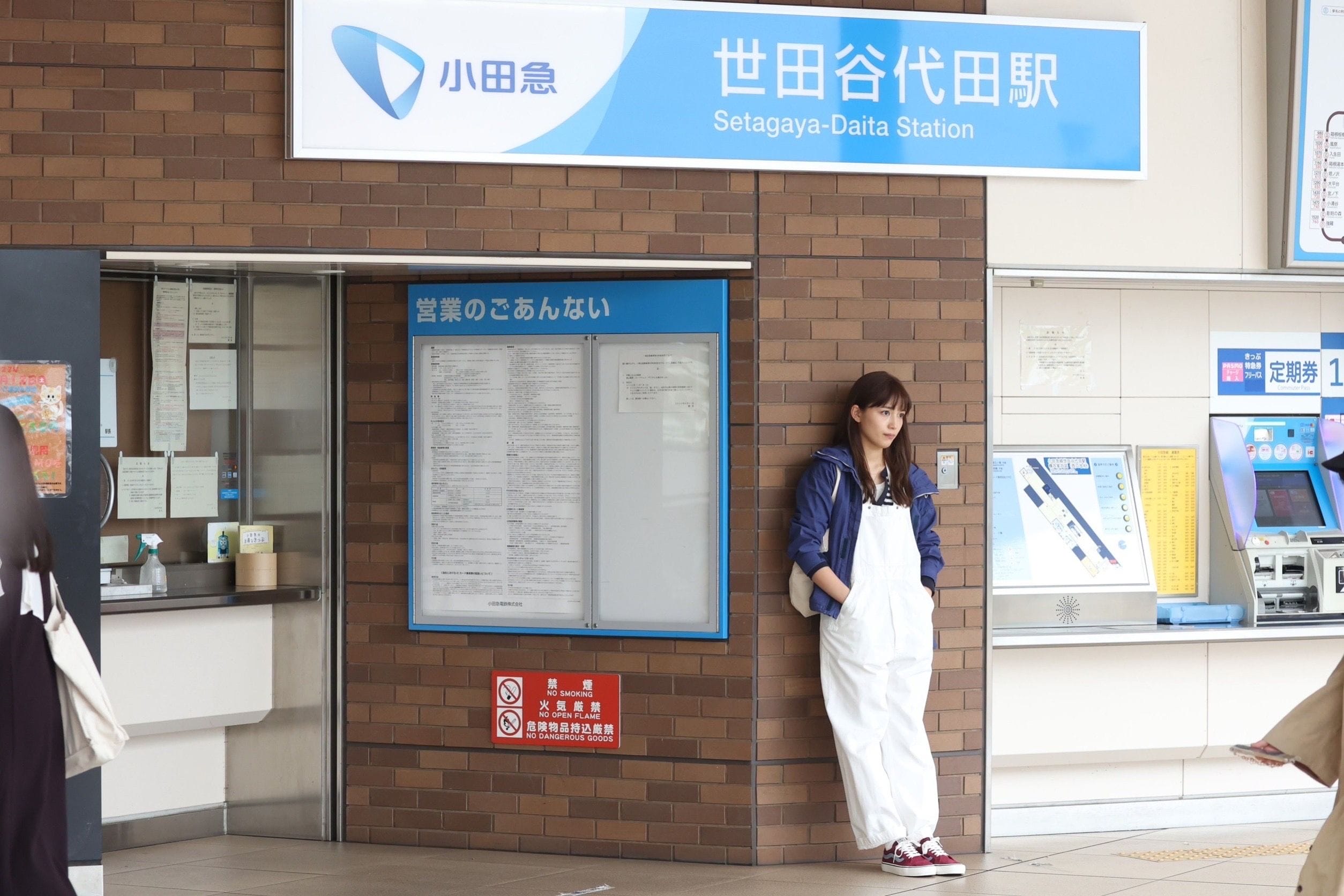 世田谷代田駅の切符売り場に立つ白いオーバーオールに青いジャケット姿の女性。.