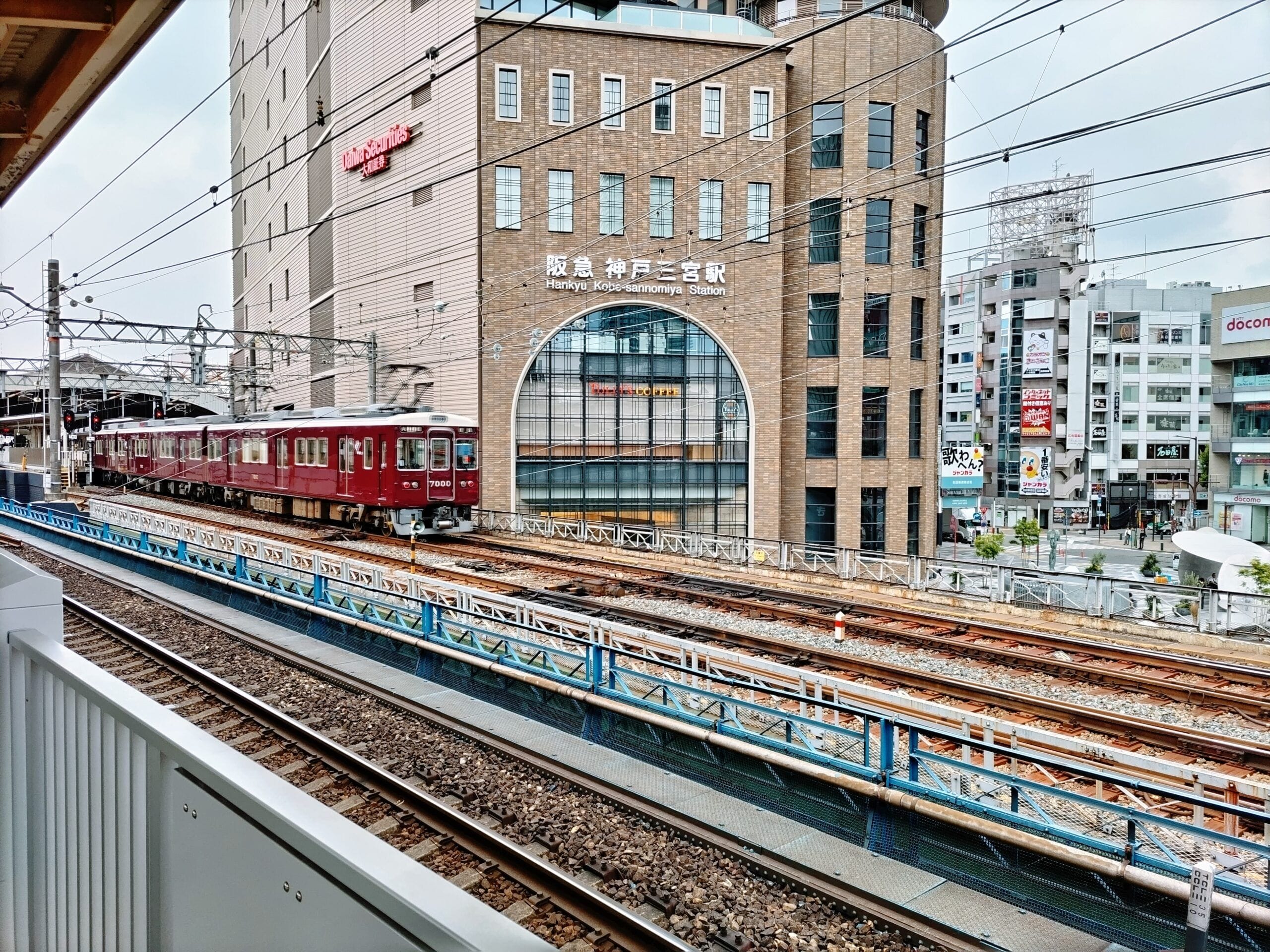 複数の商業ビルが立ち並ぶ市街地、阪急神戸三宮駅と書かれた近代的なビルの前を高架を走るマルーン色の電車。.