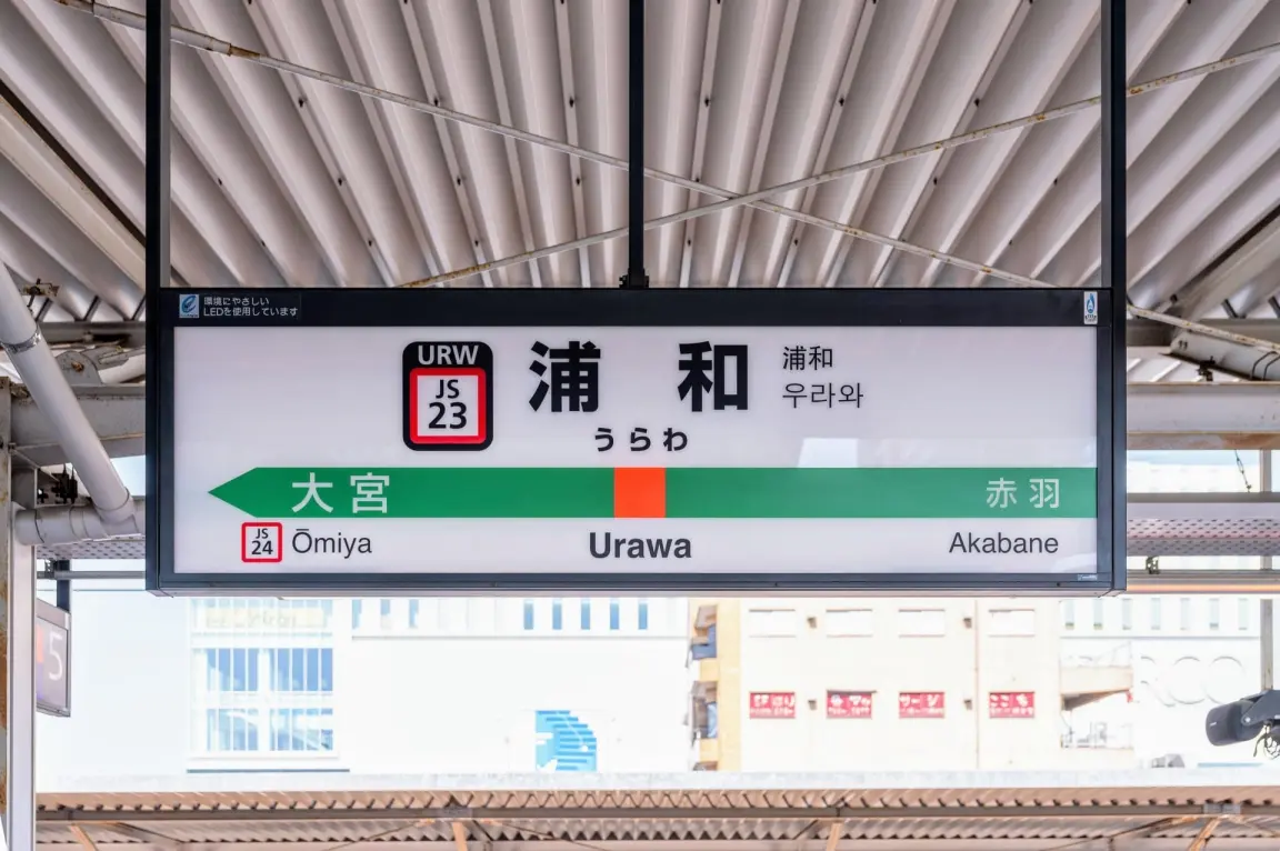 日本の浦和にある鉄道駅の看板は、金属製の屋根の下に大宮と赤羽への行き方を日本語、英語、韓国語で表示している。.