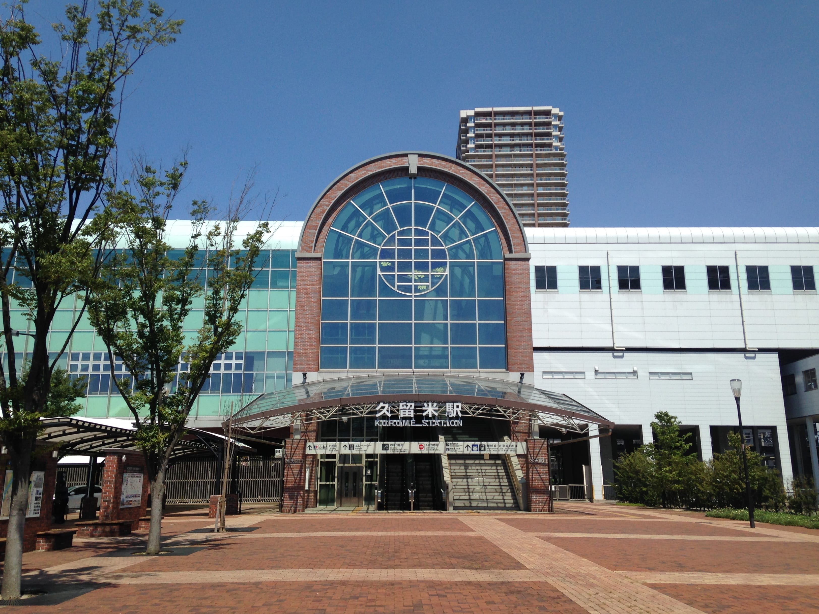 大きなアーチ型のガラス窓、レンガと白を基調としたファサード、そして澄み渡った青空の下、並木道が伸びる広場が特徴の、甲子園口駅の外観。.