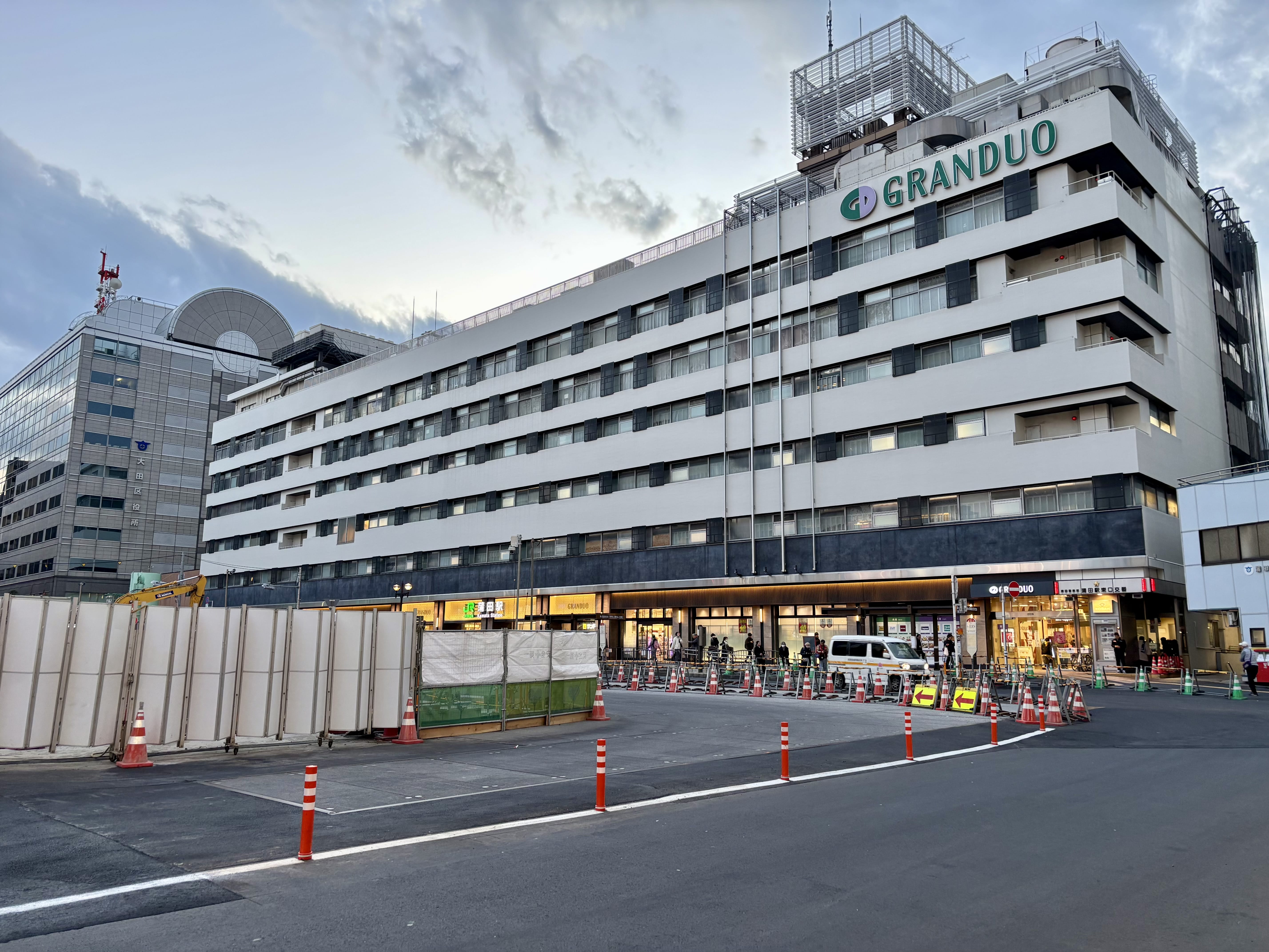 GRANDUO（グランデュオ）」と書かれた大きな立体ビルがあり、1階には店舗、工事用のバリア、コーン、前面には舗装された道路がある。.