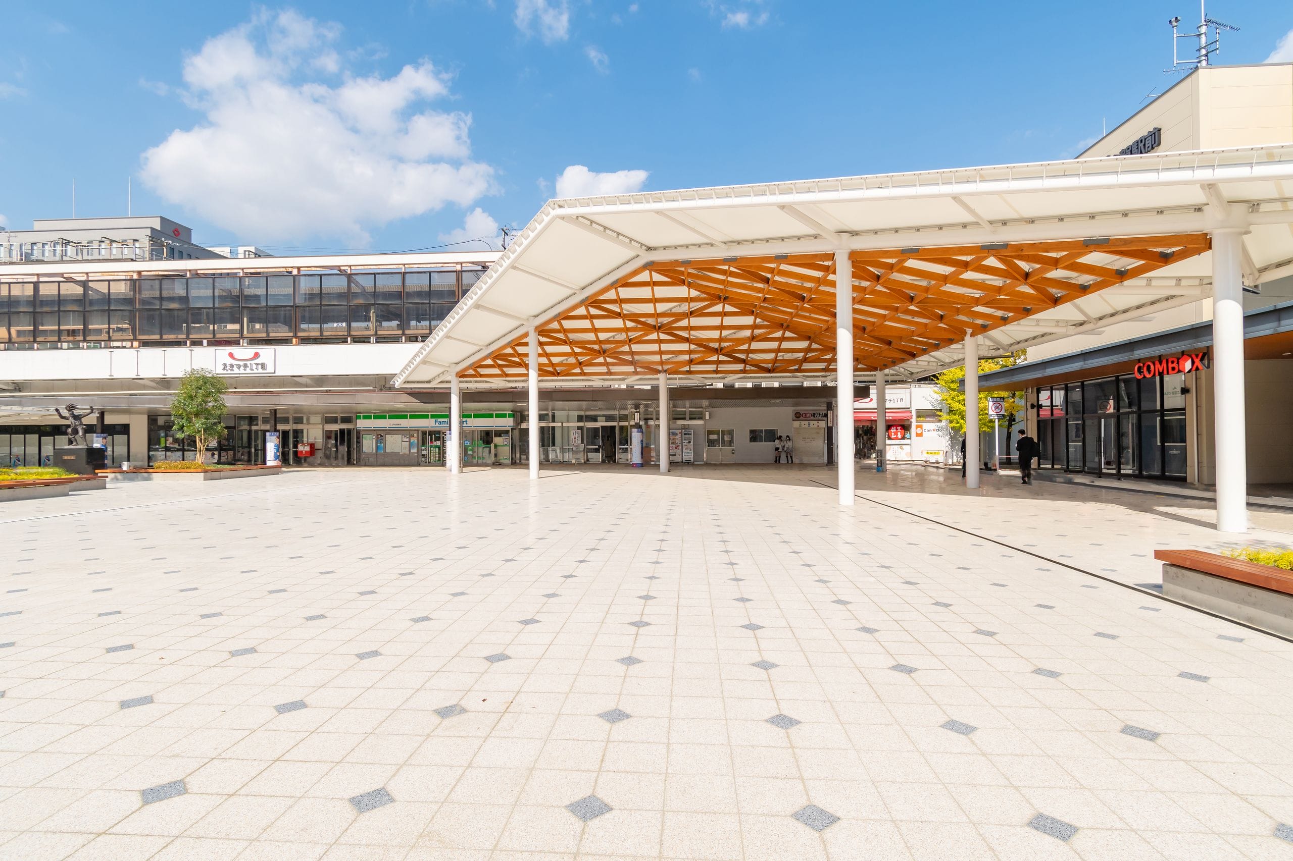 青空の下、タイル張りの床、大きなモダンなキャノピー構造、そして背景にはいくつかの店舗が並ぶ、広々とした人影のない広場。.