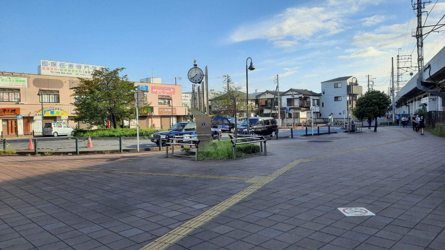 時計、街灯、黄色い触感の舗装された歩道、建物、木々、車、線路が、雲が散らばる青空の下に見える。.