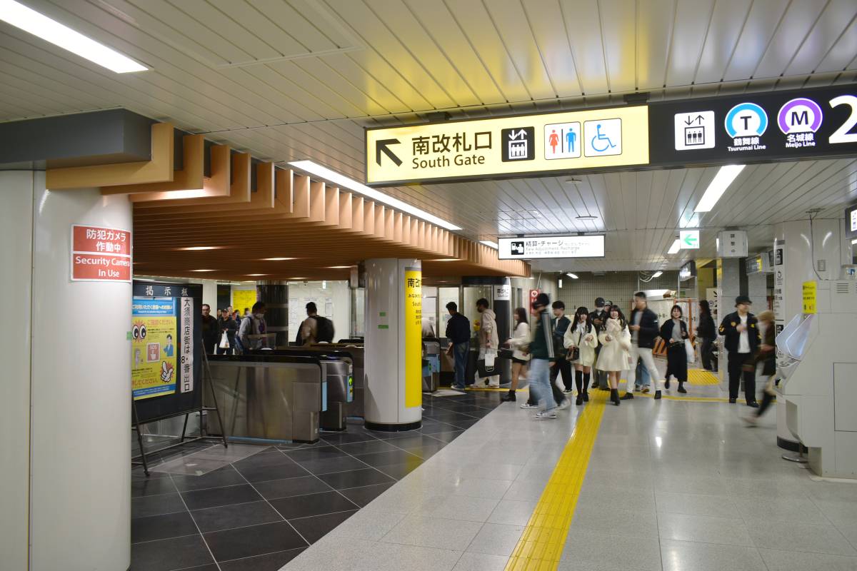 人々が駅の南口エリアを歩いている。頭上には案内表示があり、改札口や防犯カメラの警告表示が見える。.