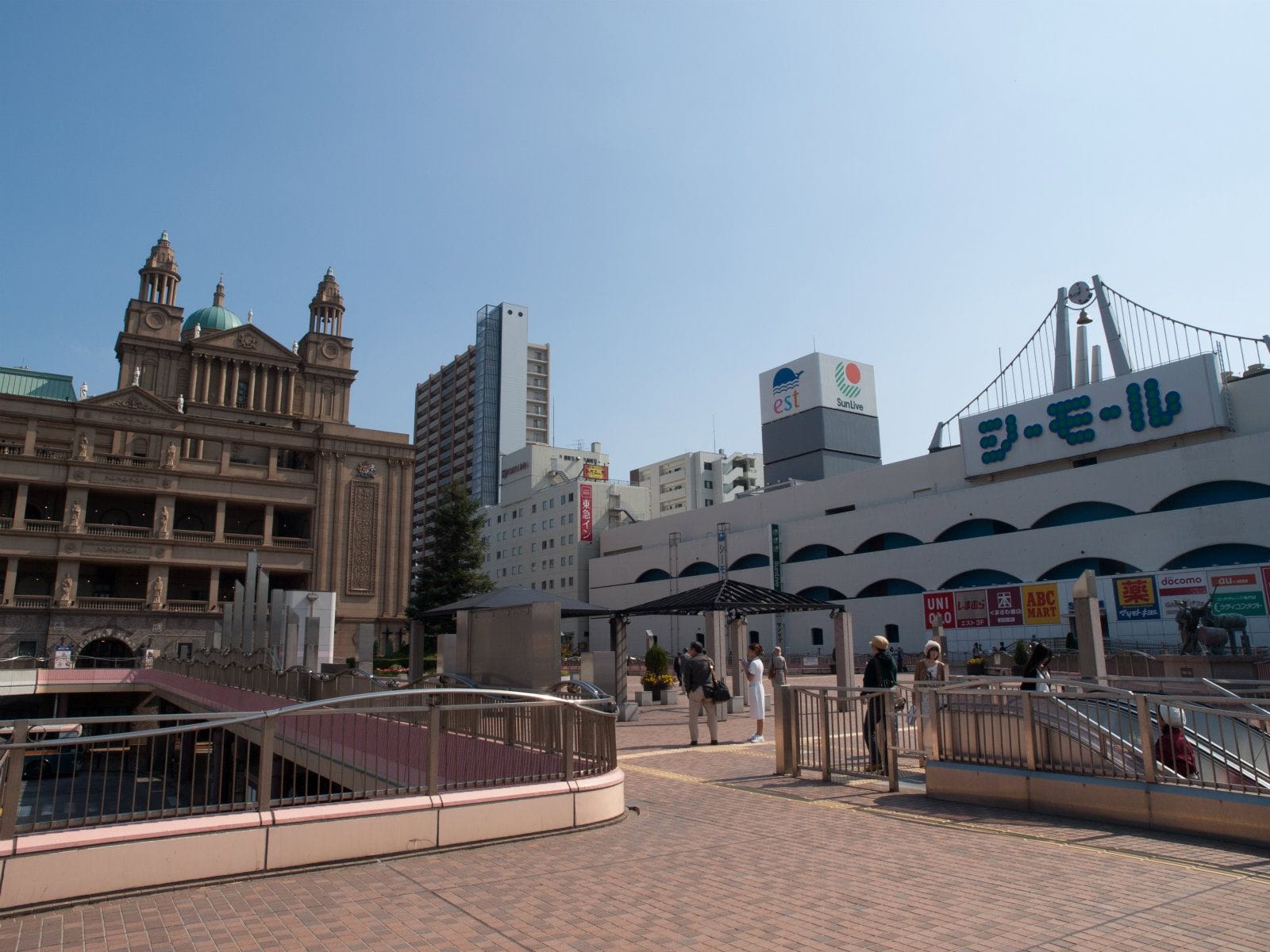青空の下、西洋風と現代的な建物が混在する都市の広場を人々が歩いている。いくつかの建物には日本語の看板が見える。.