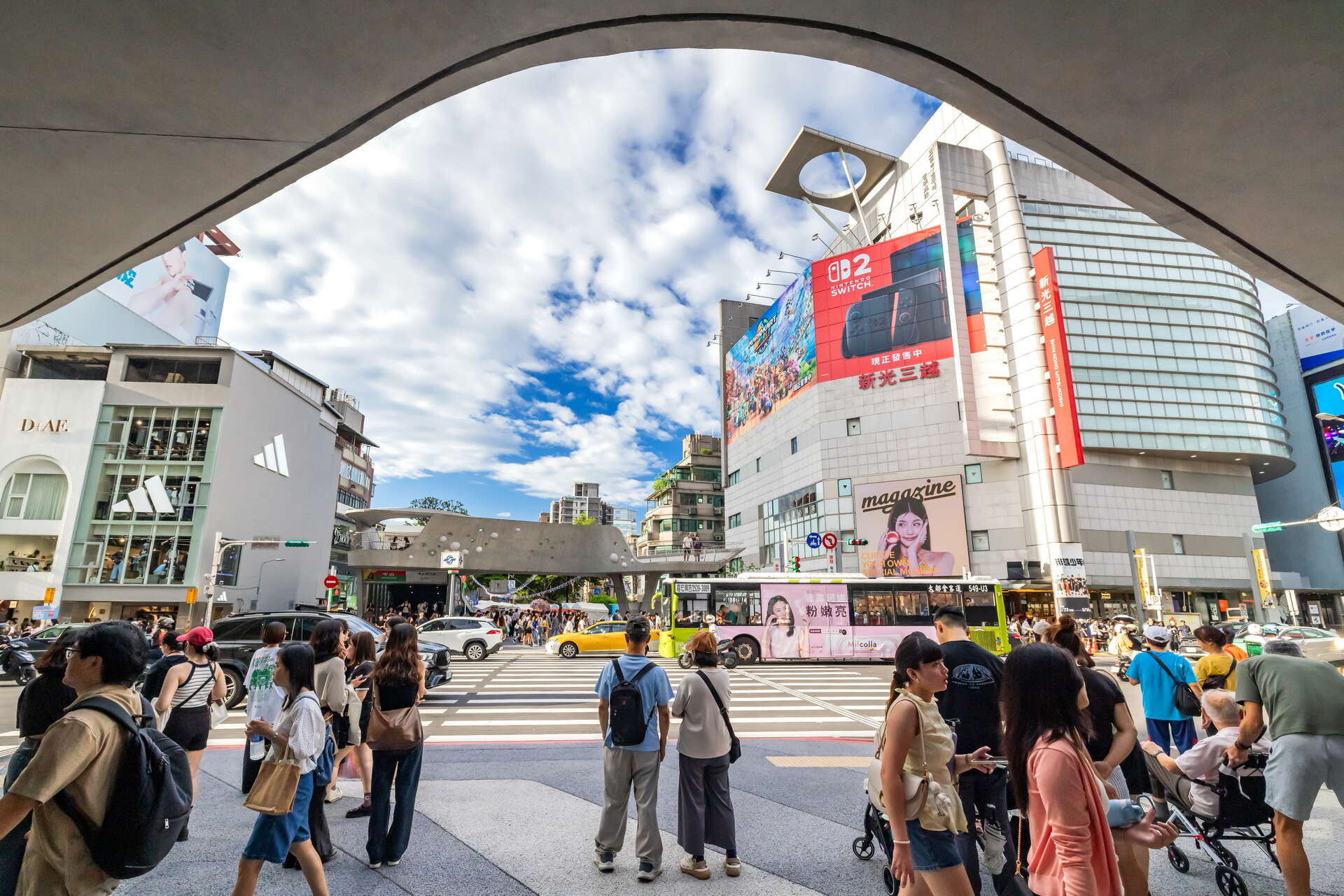 高層ビルやデジタル看板が立ち並び、背景には曇り空が広がる賑やかな街の交差点で、人々が歩き、集まっている。.