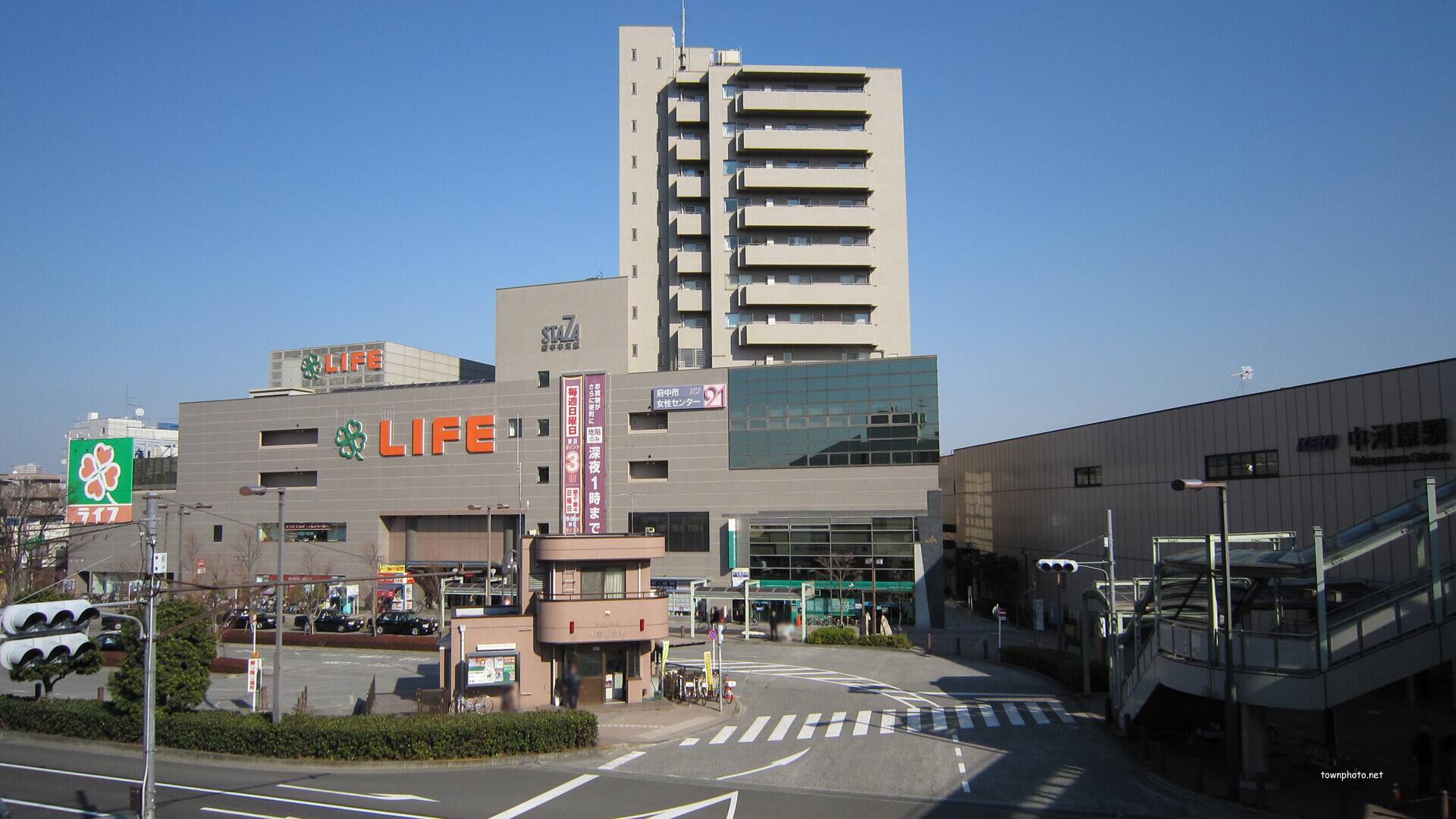青空の下、他の建物や歩行者用跨線橋に隣接して建つ、「LIFE」の看板が掲げられた高層商業ビル。.