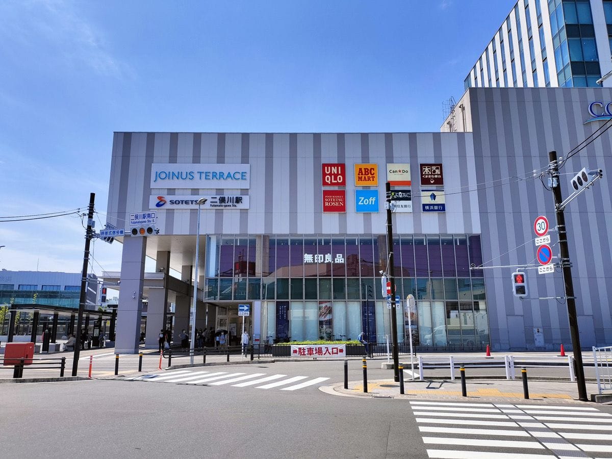 交差点のそば、澄み渡った青空の下に、「JOINUS TERRACE」と書かれたモダンなビルが立ち、その周囲にはユニクロやABCマートなど、さまざまな店舗の看板が並んでいる。.
