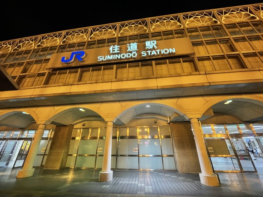 夜間の住之戸駅の外観。ライトアップされたJRの看板と、アーチ型の入口の柱が見える。.