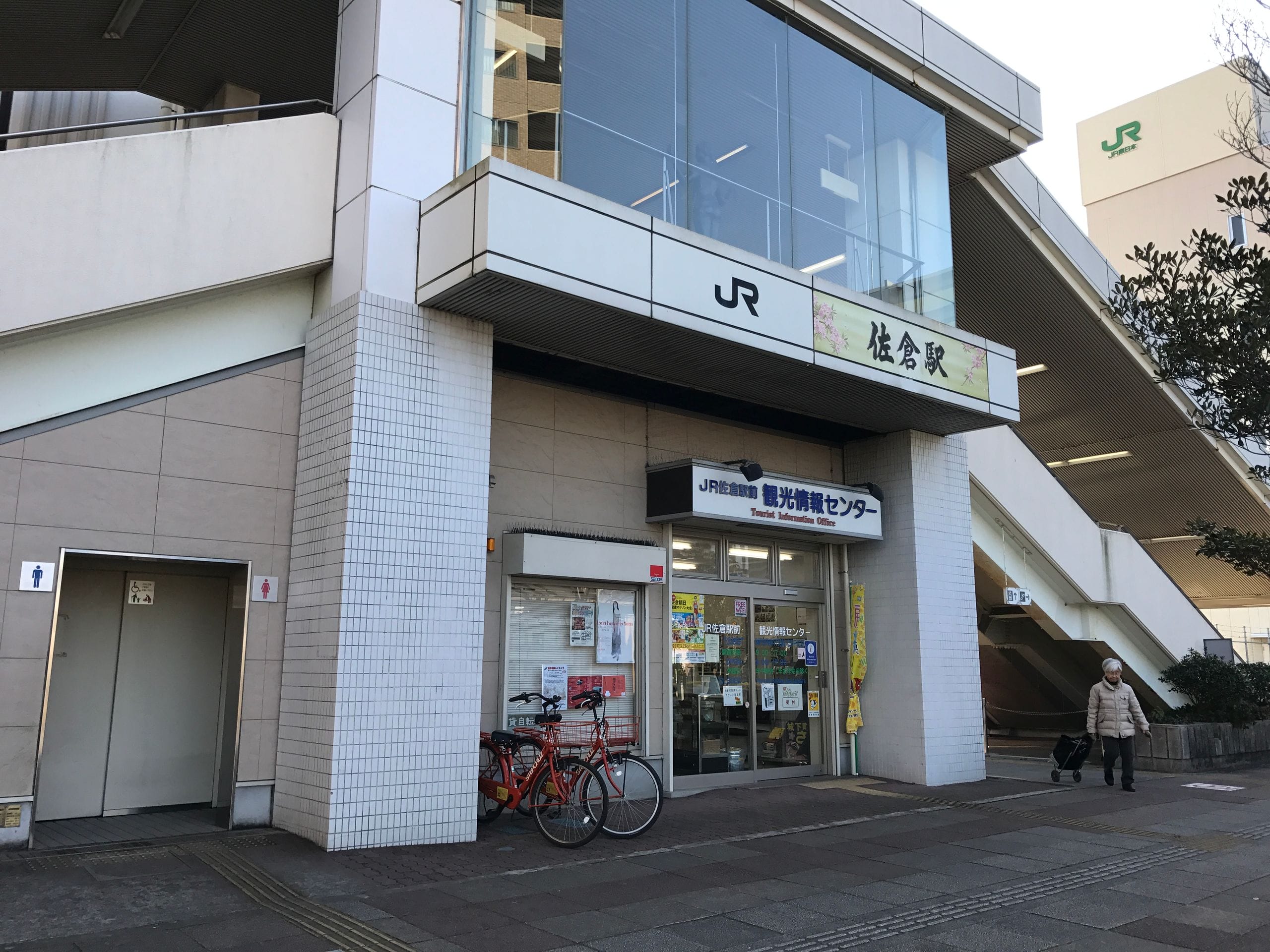 階段のあるJR駅の駅舎の外観。駅舎の外には自転車が停まっており、スーツケースを持った人が通り過ぎている。駅舎には看板や窓が見える。.