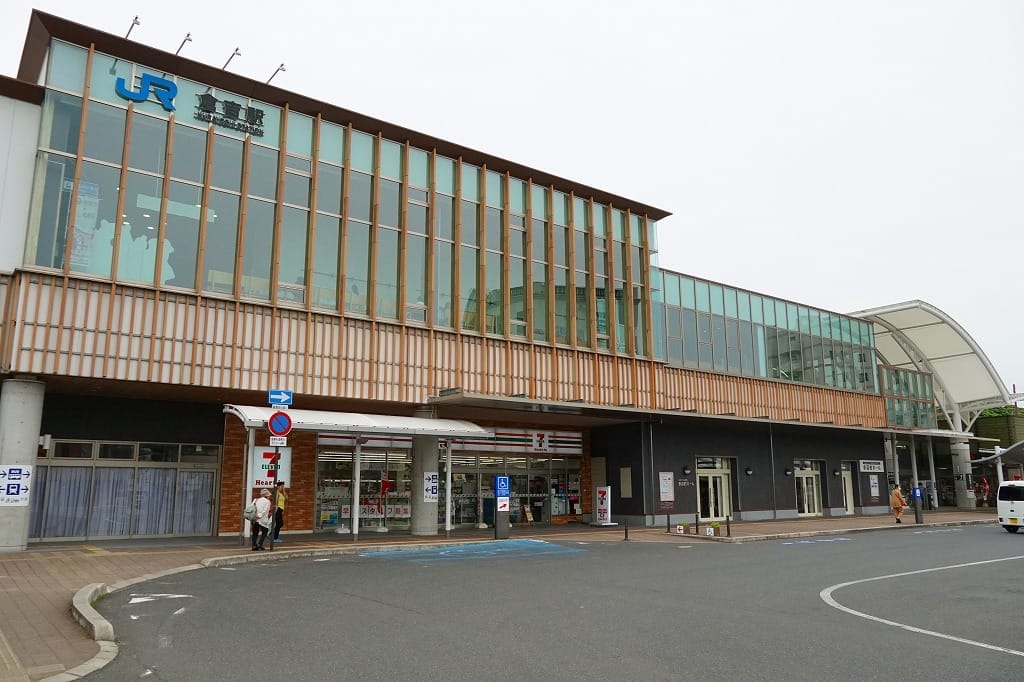 大きなガラス窓があり、複数の入口を備え、正面には屋根付きのバス停があるモダンな駅舎。その周辺を数人の人々が歩いている。.