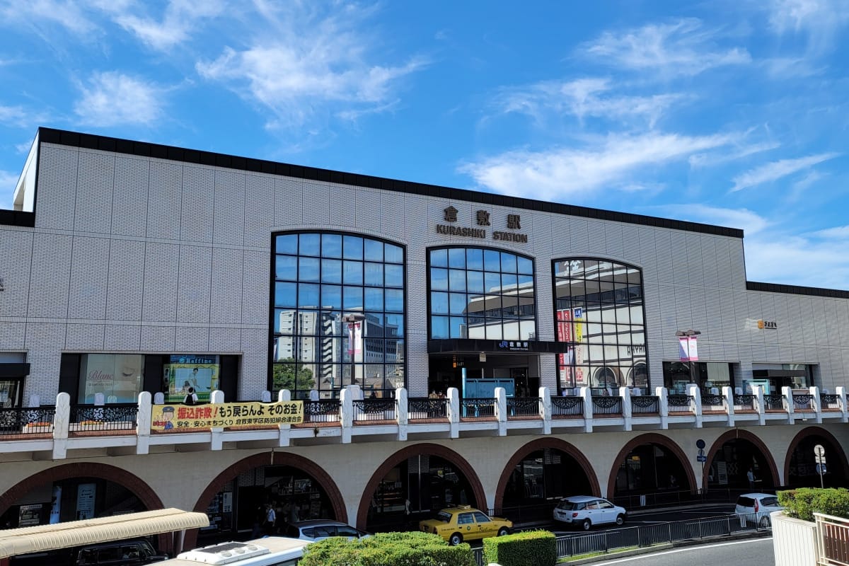 青空の下、アーチ型の入り口と大きな窓、そして目立つ案内板を備えた倉敷駅の駅舎。その前には、黄色いタクシーを含む車が停まっている。.