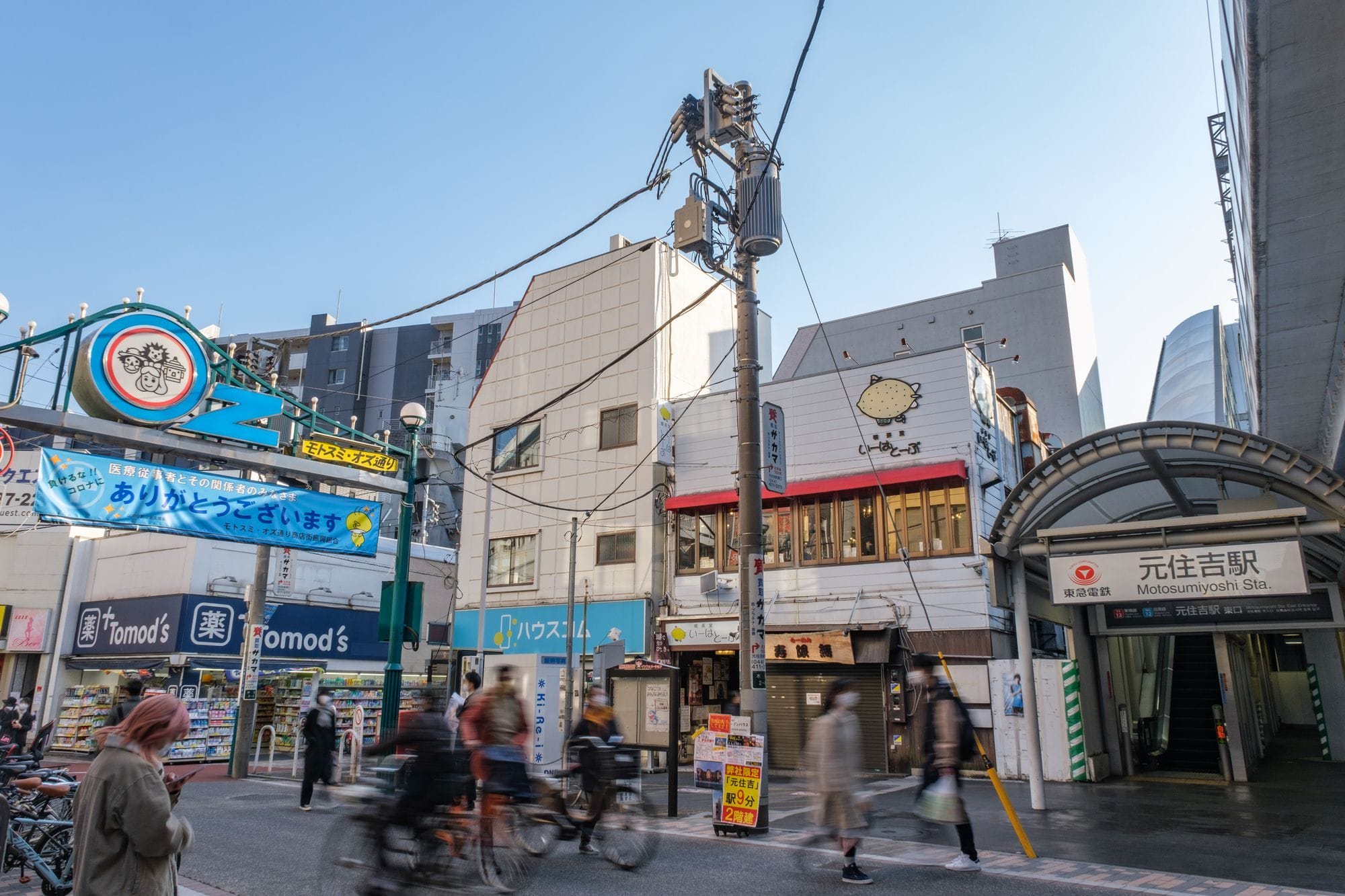 日本の元向山駅前の賑やかな街並み。澄み渡った空の下、歩行者や自転車、お店、そして背景には高層ビルが並んでいる。.