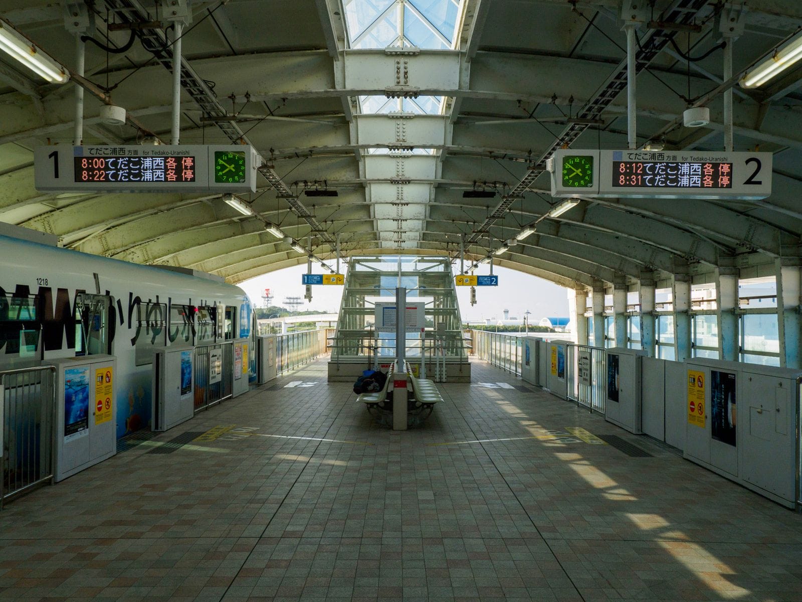 2本の線路、デジタル時刻表、中央のベンチ、ガラス屋根の下に停車中の列車。.