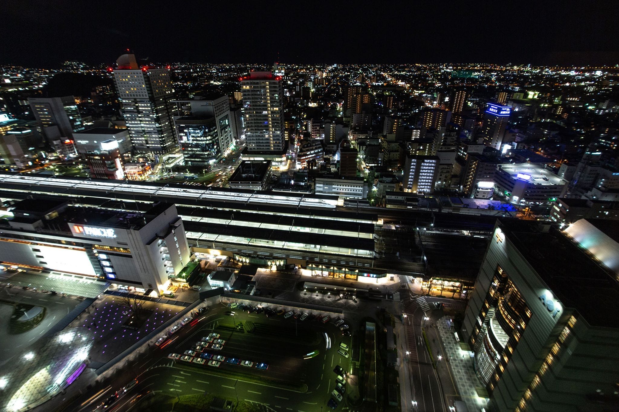 夜間の街の空撮写真。ライトアップされたビル、駅、駐車中の車、そして街灯の明かりが遠くまで続く賑やかな通りが写っている。.