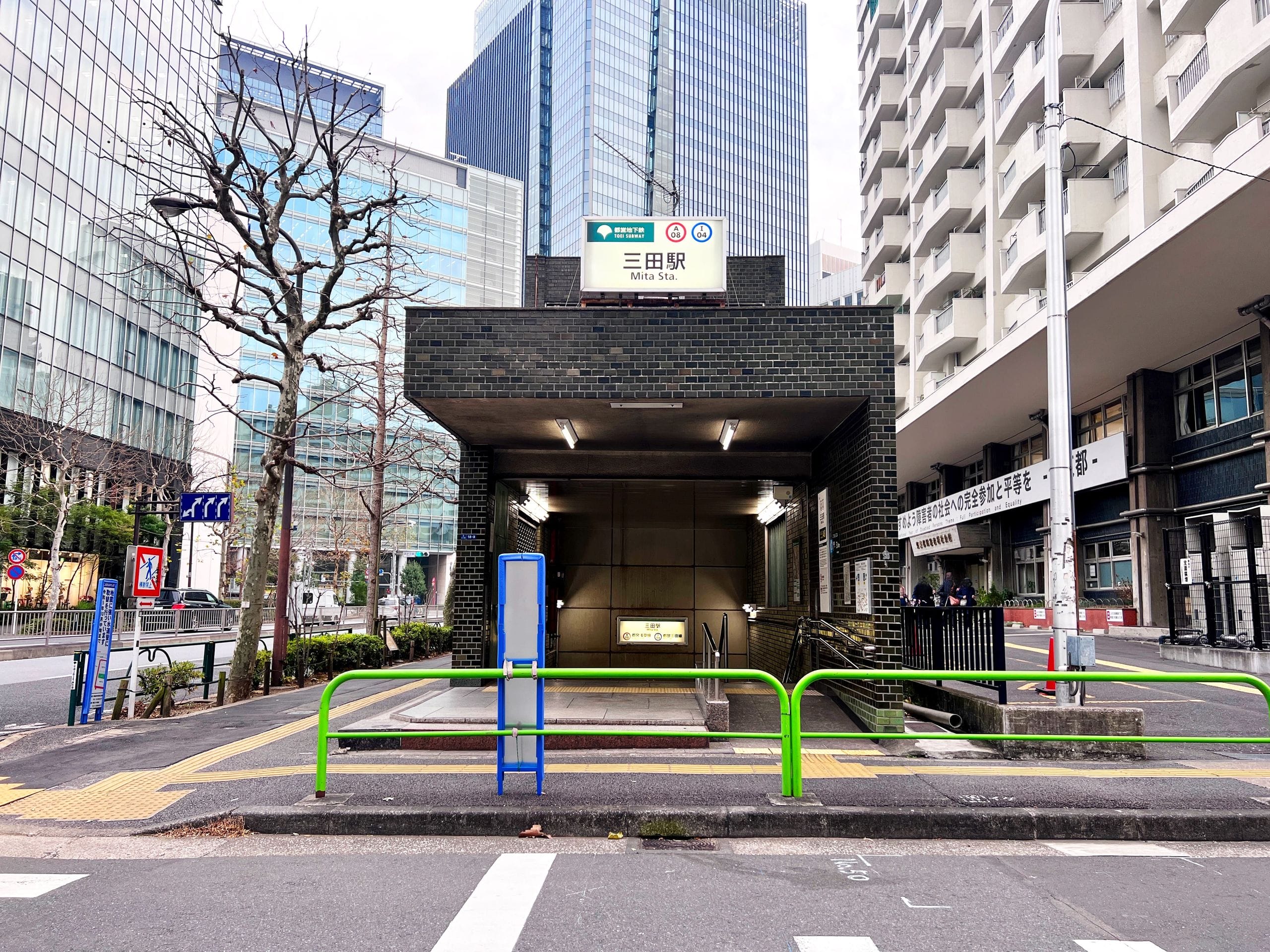 東京・三田駅の入り口。周囲には近代的なビルが立ち並び、手前には看板や木、緑色の手すりが写っている。.