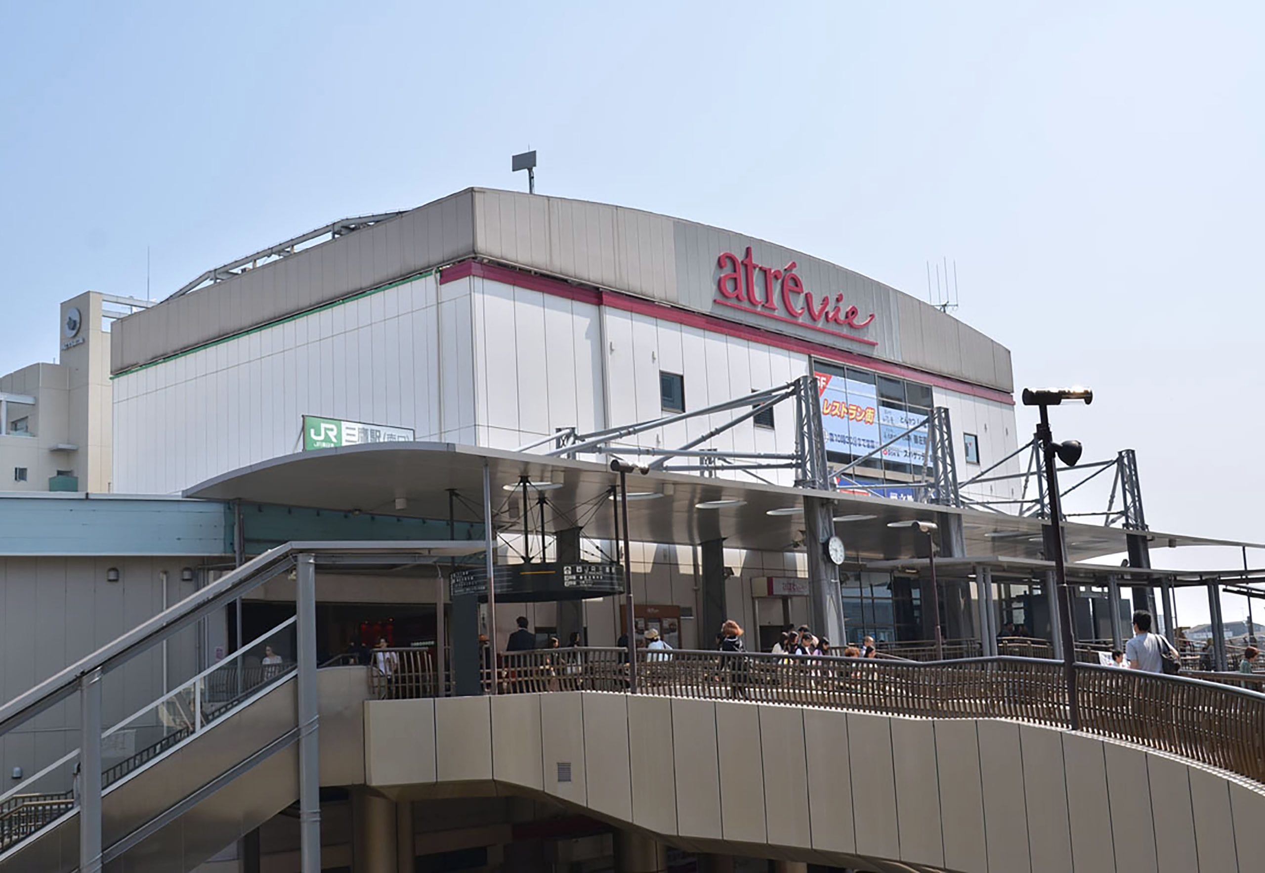 晴れ渡った空の下、高架歩道を歩く人々の姿と、「アトレ・ヴィ」の看板、JR東日本の看板が掲げられたモダンな建物の外観。.